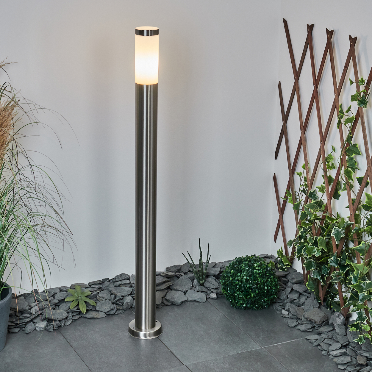 Eine beleuchtete, metallische Außenlampe steht vor einer weißen Wand. Grüne Pflanzen und Schiefersteine umgeben die Lampe. Rechts ist ein hölzernes Spalier.