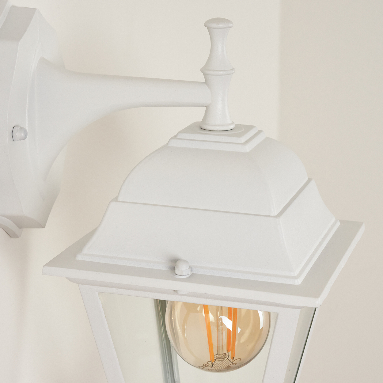Eine weiße, an der Wand montierte Außenlampe mit Glasfront und sichtbarer Glühbirne. Die Lampe ist an einer weißen Wand befestigt.