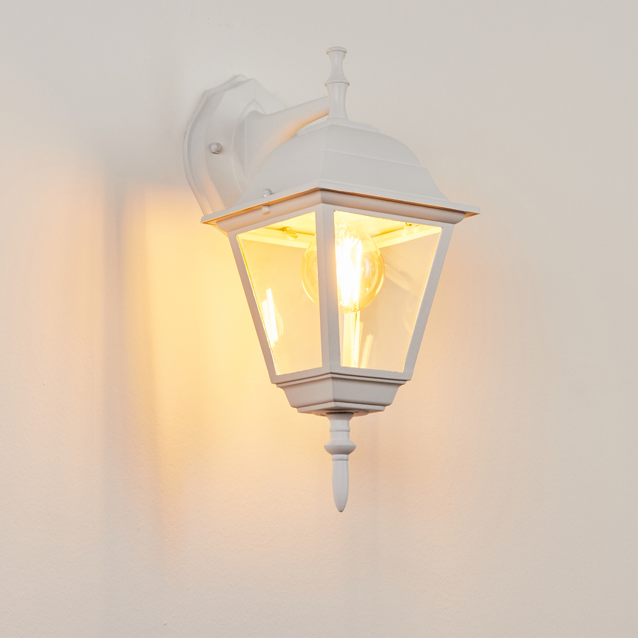 Eine weiße Außenlampe mit einer brennenden Glühbirne hängt an einer hellen Wand. Die Lampe hat ein klassisches Design mit Glaspaneelen und einer dekorativen Spitze.