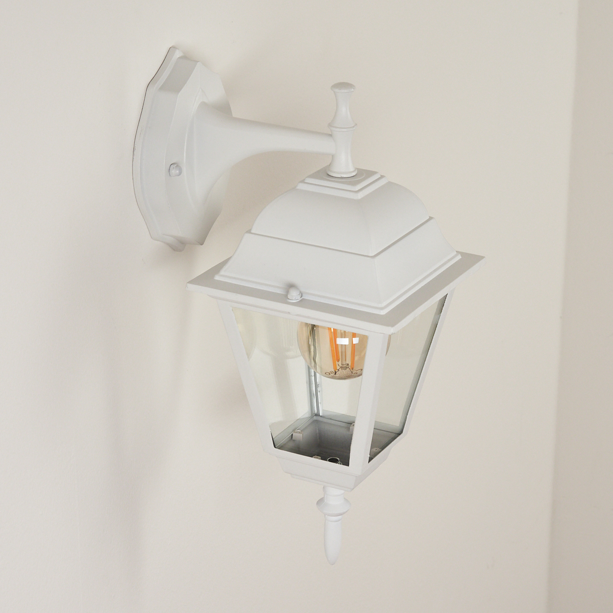 Eine weiße, an der Wand montierte Außenlampe mit Glasscheiben. Im Inneren befindet sich eine orangefarbene Glühbirne. Sie befindet sich an einer weißen Wand.
