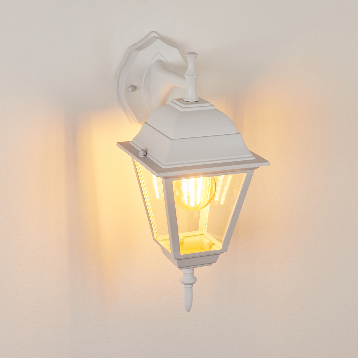Eine weiße, verzierte Außenlampe ist an einer Wand befestigt. Die Lampe hat ein Glasgehäuse mit einer leuchtenden Glühbirne.