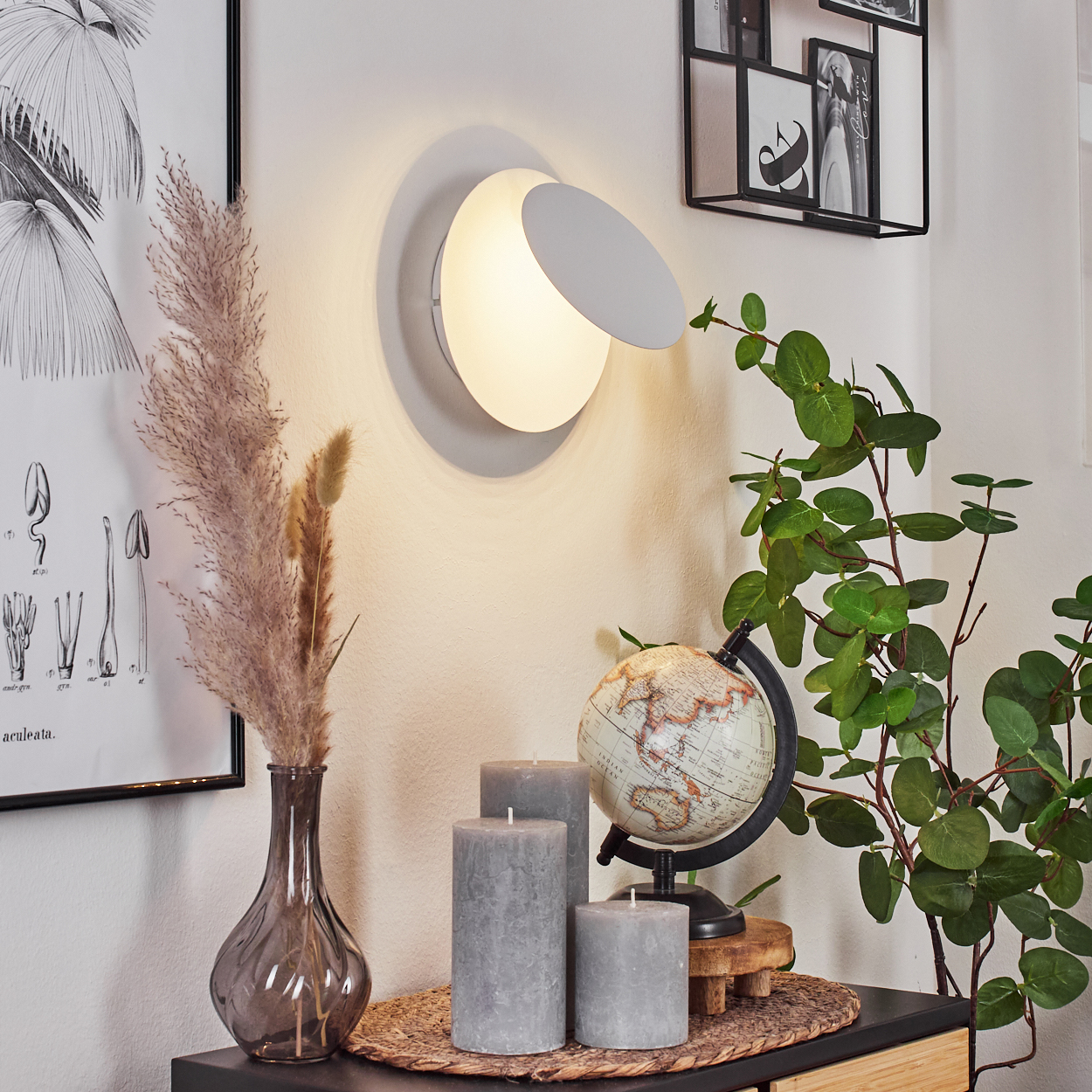 Eine gut beleuchtete Innenszene mit Wandlampe, Pflanzendekoration, Kerzen und Globus auf einem Tisch. Die Einrichtung ist schlicht und elegant.