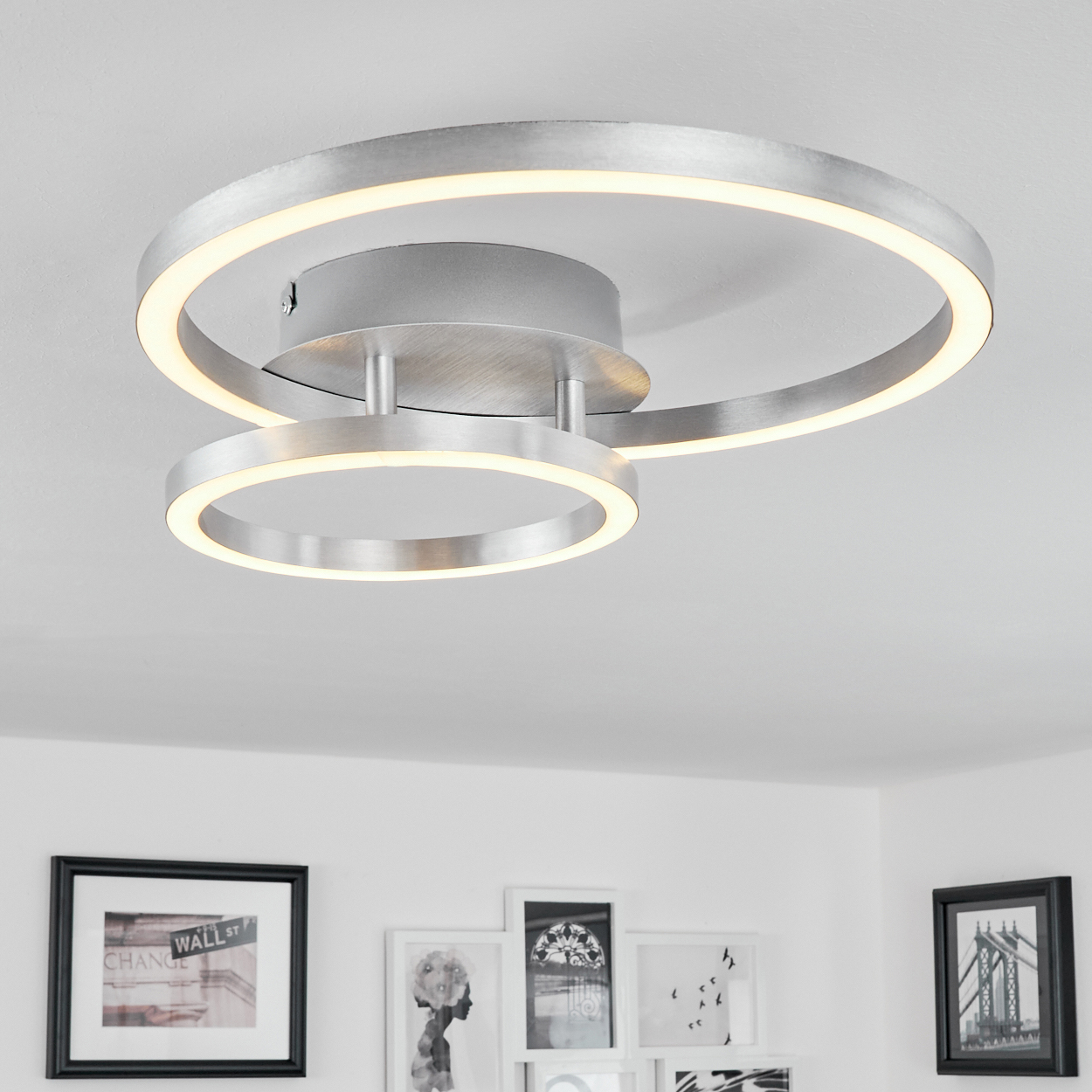 Moderne silberne Deckenleuchte mit zwei beleuchteten Ringen. Darunter hängen gerahmte Bilder an einer weißen Wand.