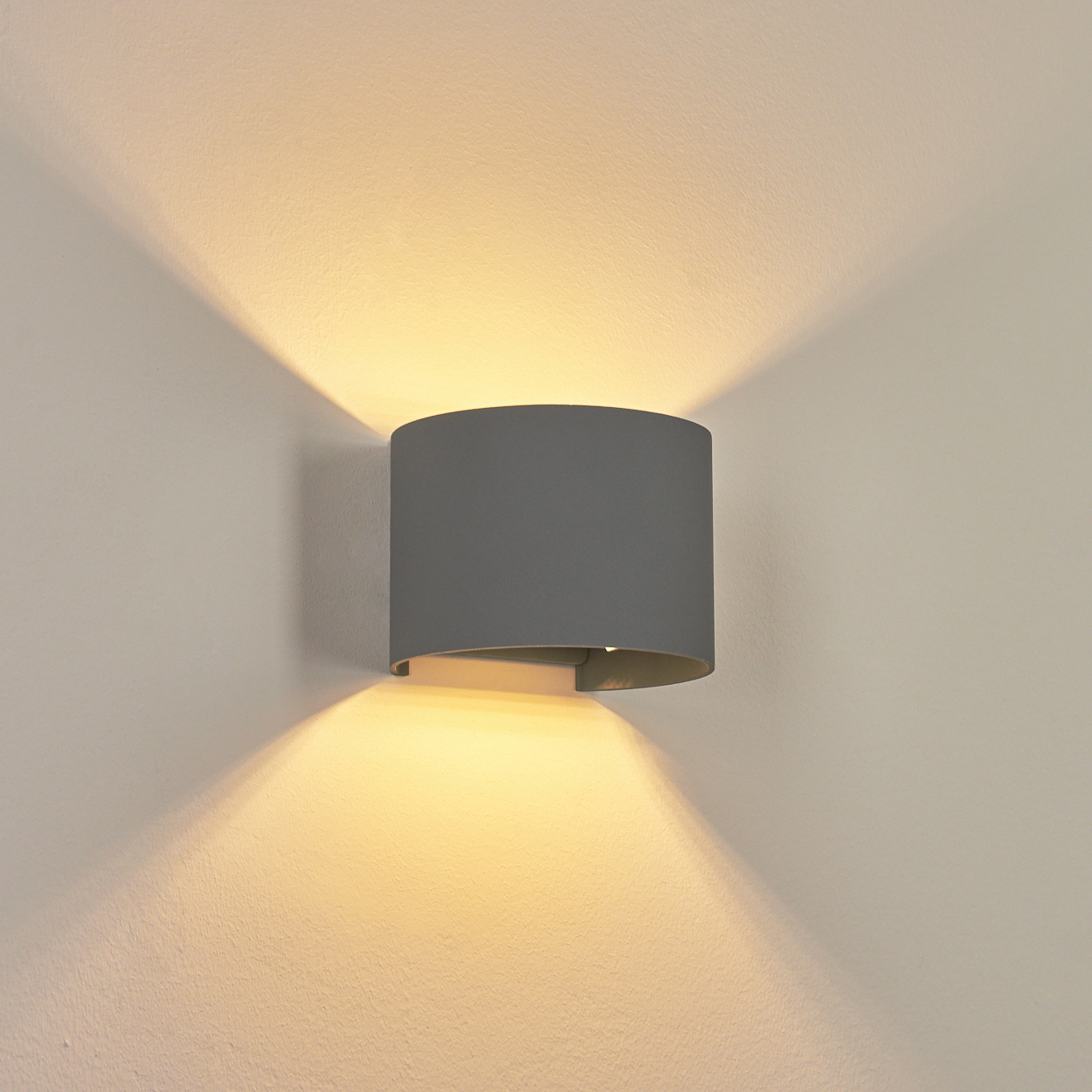 Eine graue Wandleuchte ist an einer hellen Wand angebracht. Die Lampe strahlt Licht nach oben und unten aus und erzeugt eine helle Beleuchtung.