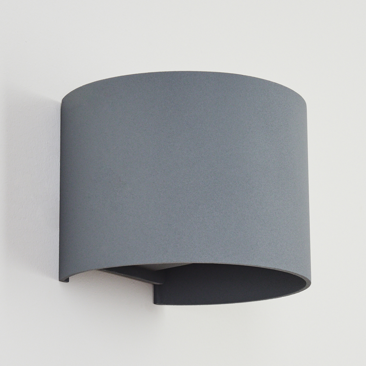 Eine graue, halb zylindrische Wandleuchte an einer weißen Wand. Die Lampe hat ein gebogenes Design und eine dunkle Unterseite.