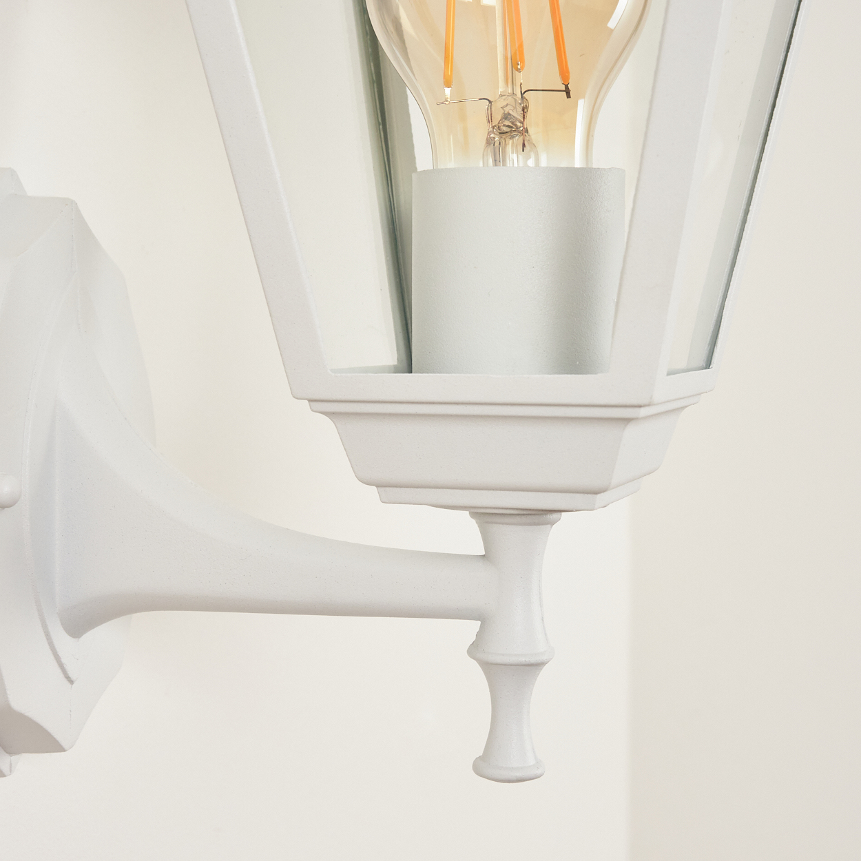 Weiße Außenlampe mit Glasscheiben und sichtbarer Glühbirne. Die Lampe ist an einer weißen Wand montiert, Detailansicht.