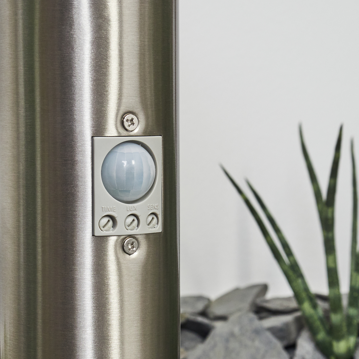 Eine Metallstange mit Sensor und Bedienfeld. Im Hintergrund eine Pflanze und Steine vor einer weißen Wand.