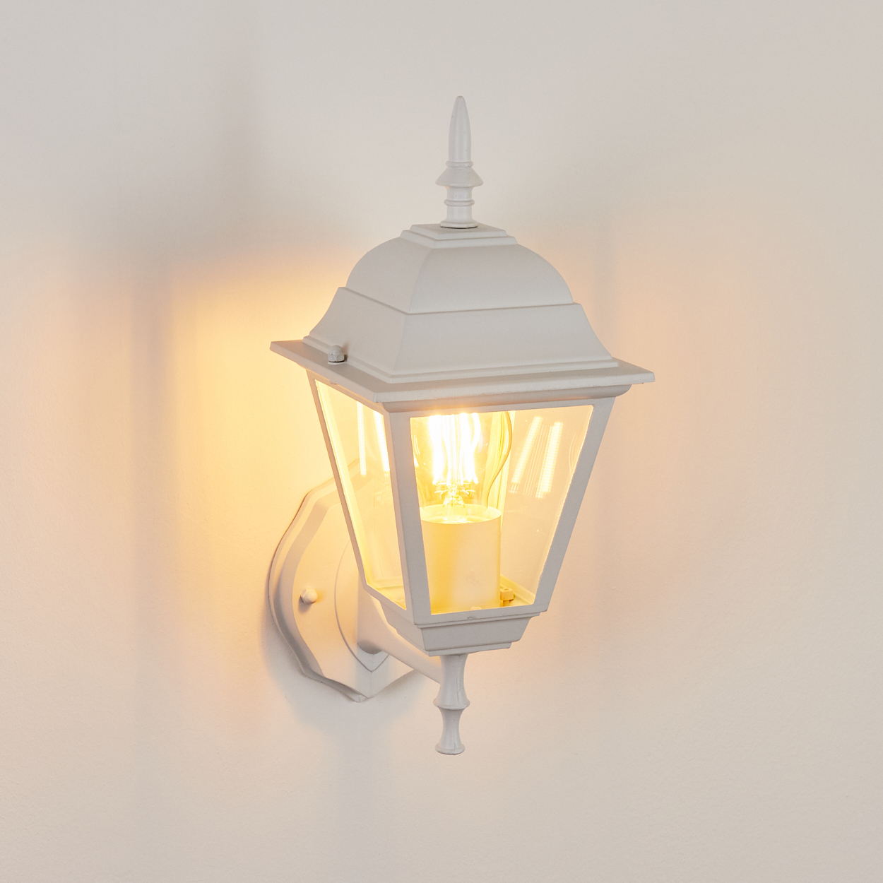 Eine weiße, an der Wand montierte Außenlampe mit einer leuchtenden Glühbirne. Die Lampe befindet sich an einer weißen Wand.
