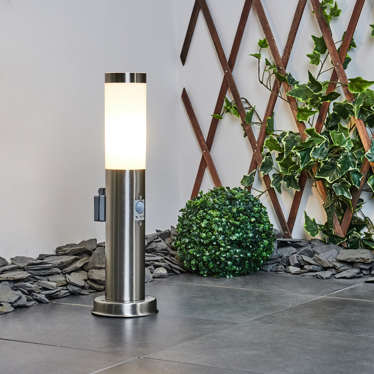 Eine silberne Außenlampe leuchtet, mit einem kleinen grünen Busch, einem hölzernen Gitter und dunklen grauen Schiefersteinen im Hintergrund.