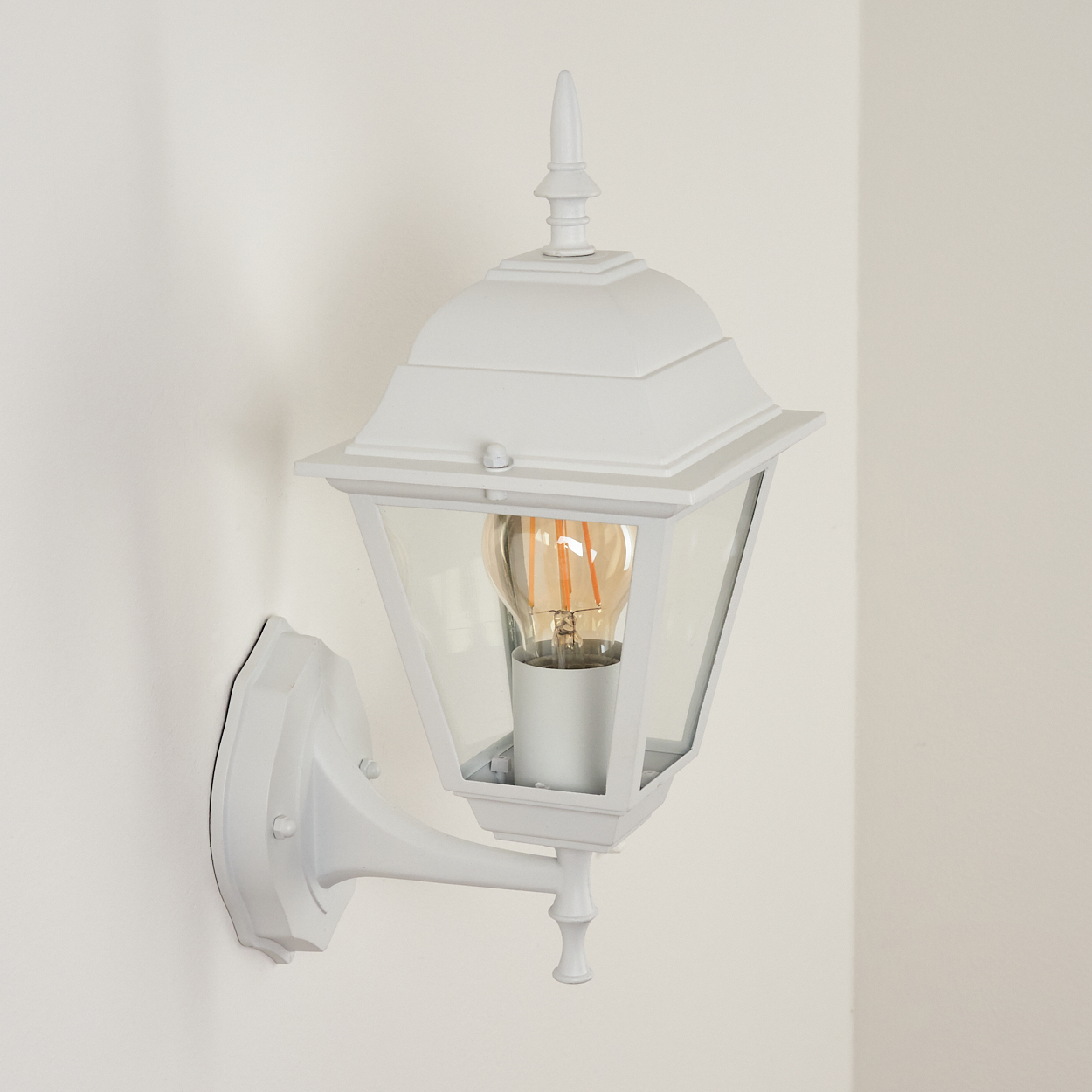 Eine weiße Wandleuchte mit Glasgehäuse, die eine Glühbirne enthält. Die Lampe hat eine dekorative Oberseite und ist an einer weißen Wand befestigt.