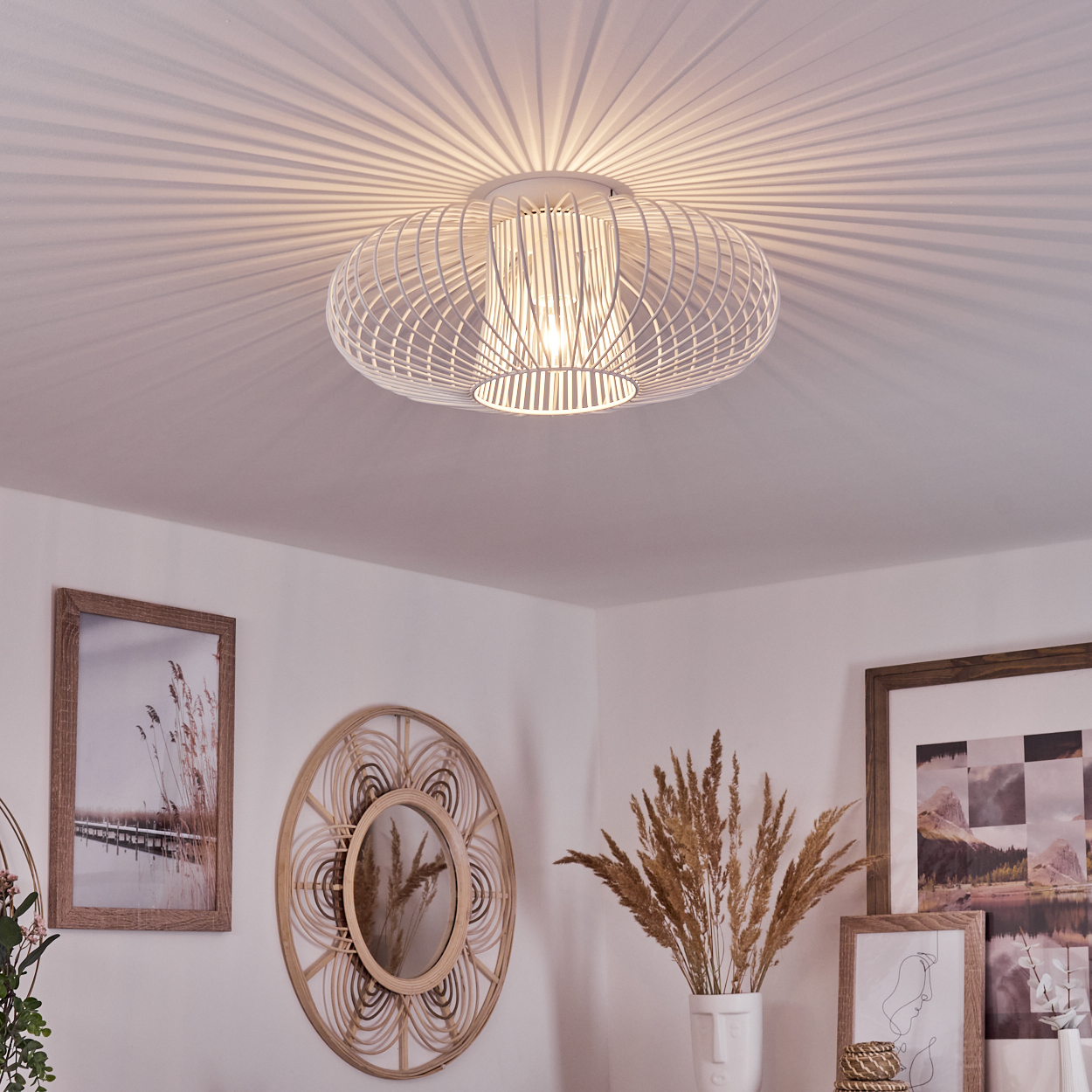 Eine weiße Deckenlampe mit kreisförmigem Käfigdesign hängt von der Decke. Kunstwerke und ein Spiegel sind an der Wand. Eine Vase mit getrockneten Pflanzen.