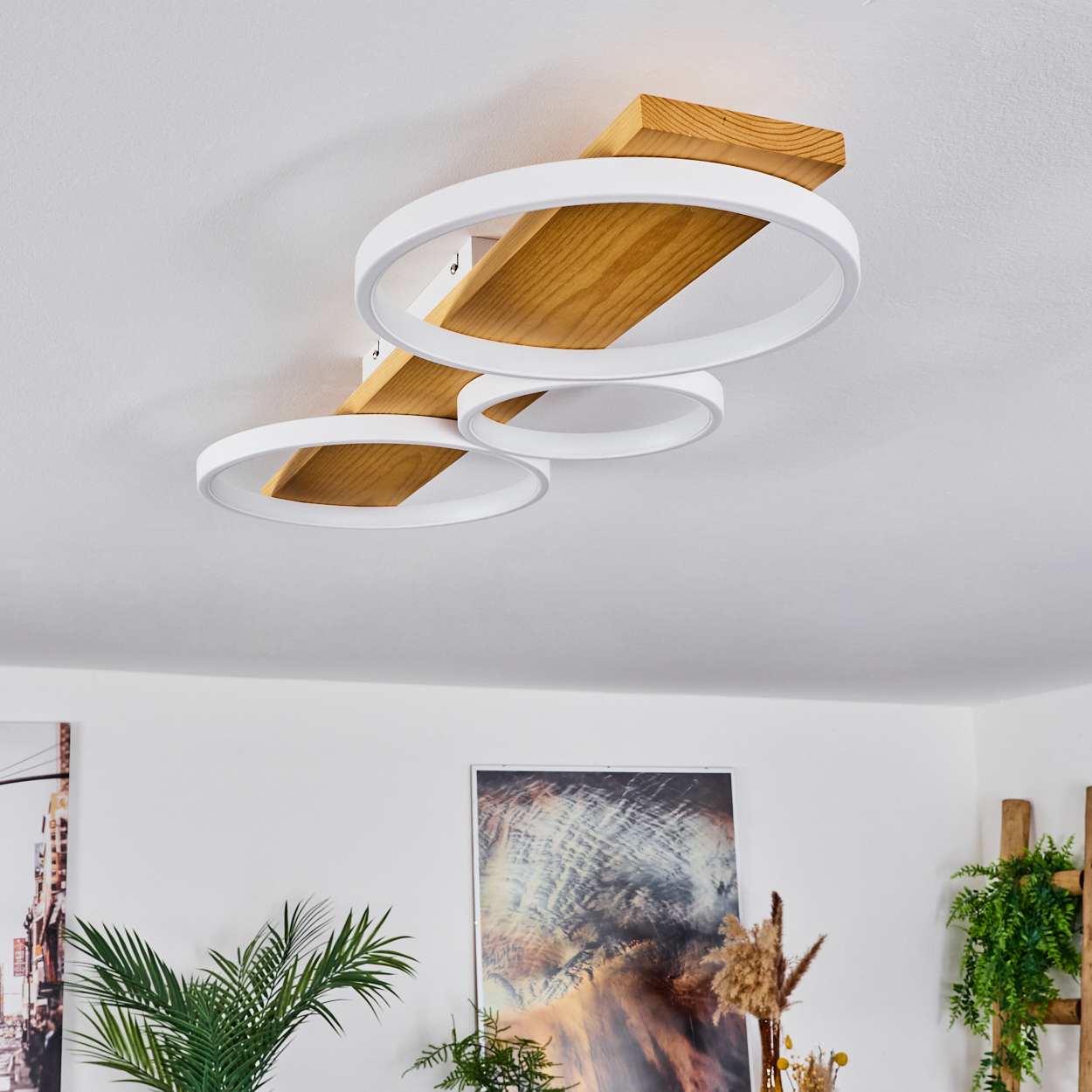 Eine moderne Deckenlampe mit Holzbalken und drei weißen kreisförmigen Lichtern. Darunter ein Raum mit Pflanzen und Bildern an den Wänden.