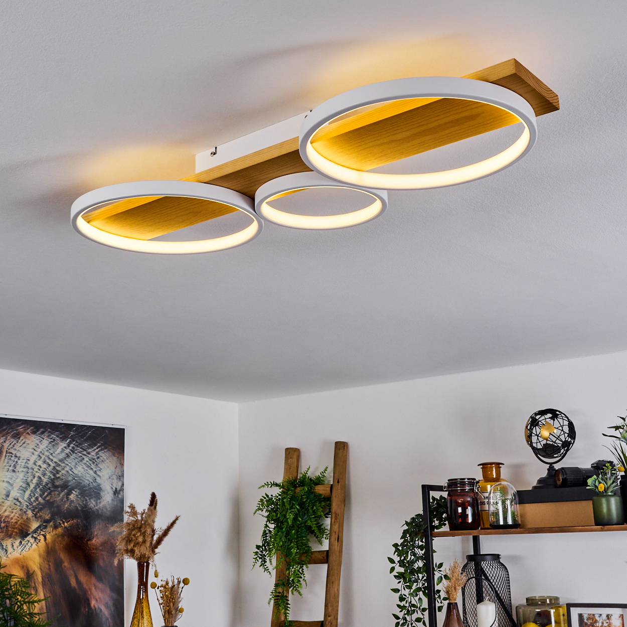 Eine moderne Deckenleuchte mit drei beleuchteten Ringen. Das Licht ist an einem Holzbalken befestigt. Darunter sind Dekorationsartikel und Pflanzen zu sehen.