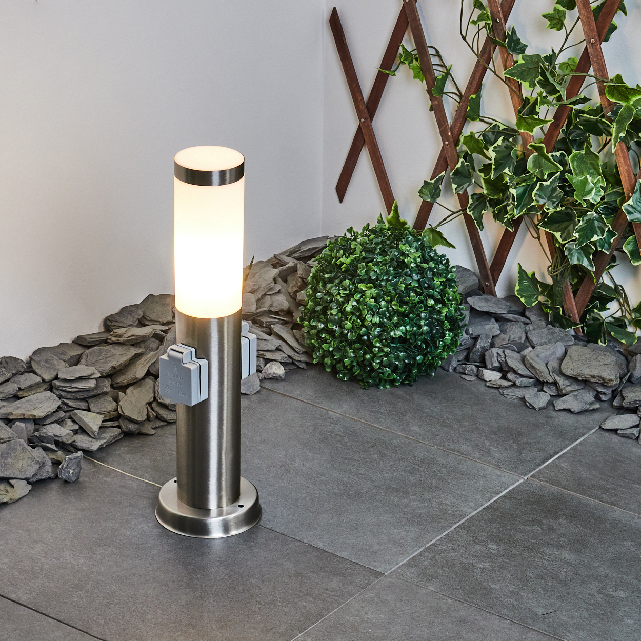 Eine zylindrische Außenlampe mit Licht, Steckdosen, umgeben von grauen Steinen, einer Hecke und einem hölzernen Gitter mit Grün.