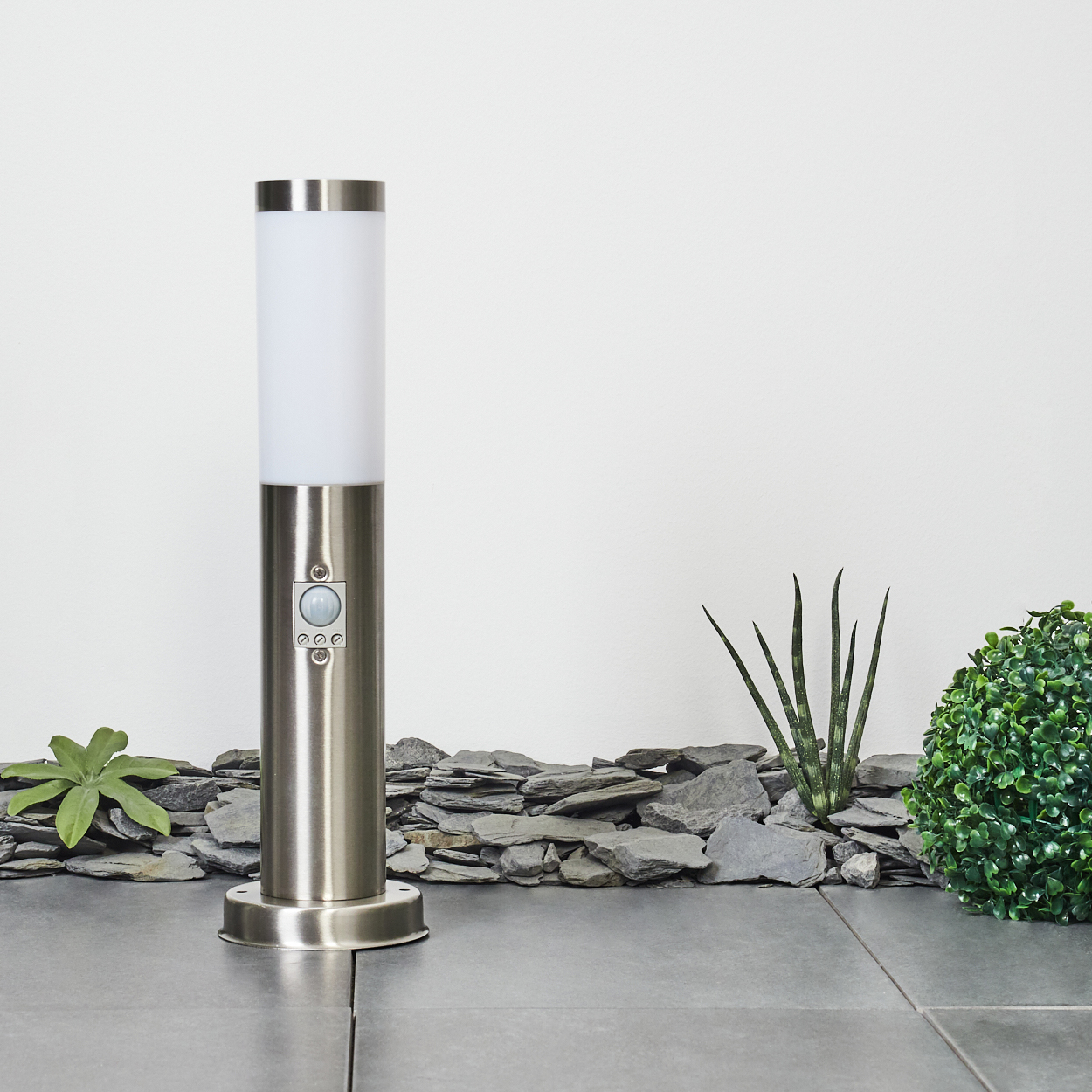 Eine Edelstahl-Lampe steht auf grauen Fliesen. Grüne Pflanzen und graue Steine ​​im Hintergrund vor einer weißen Wand.