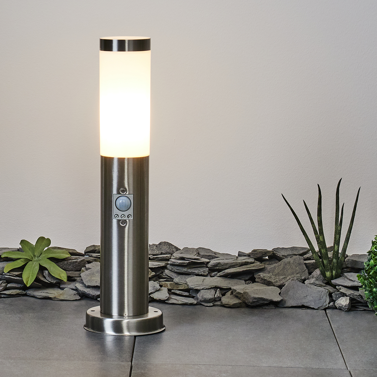 Eine zylindrische Außenlampe leuchtet. Sie hat einen Bewegungssensor. Sie steht auf einem dunkelgrauen Fliesenboden. Im Hintergrund sind Pflanzen und Steine.