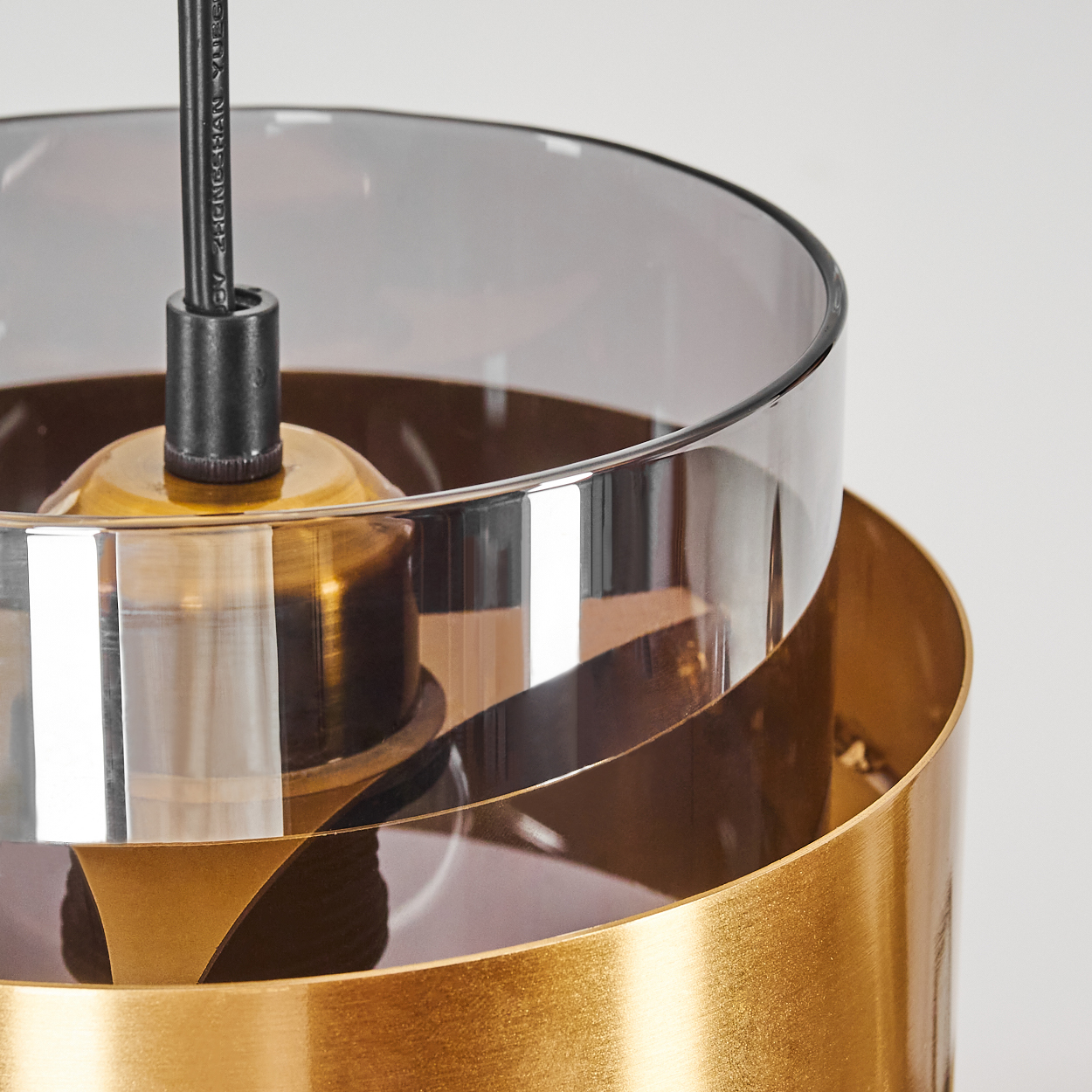 Nahaufnahme einer modernen Pendelleuchte. Sie hat einen goldenen Sockel und Rand sowie einen transparenten, rauchigen Glasschirm. Ein schwarzes Kabel hängt von oben.