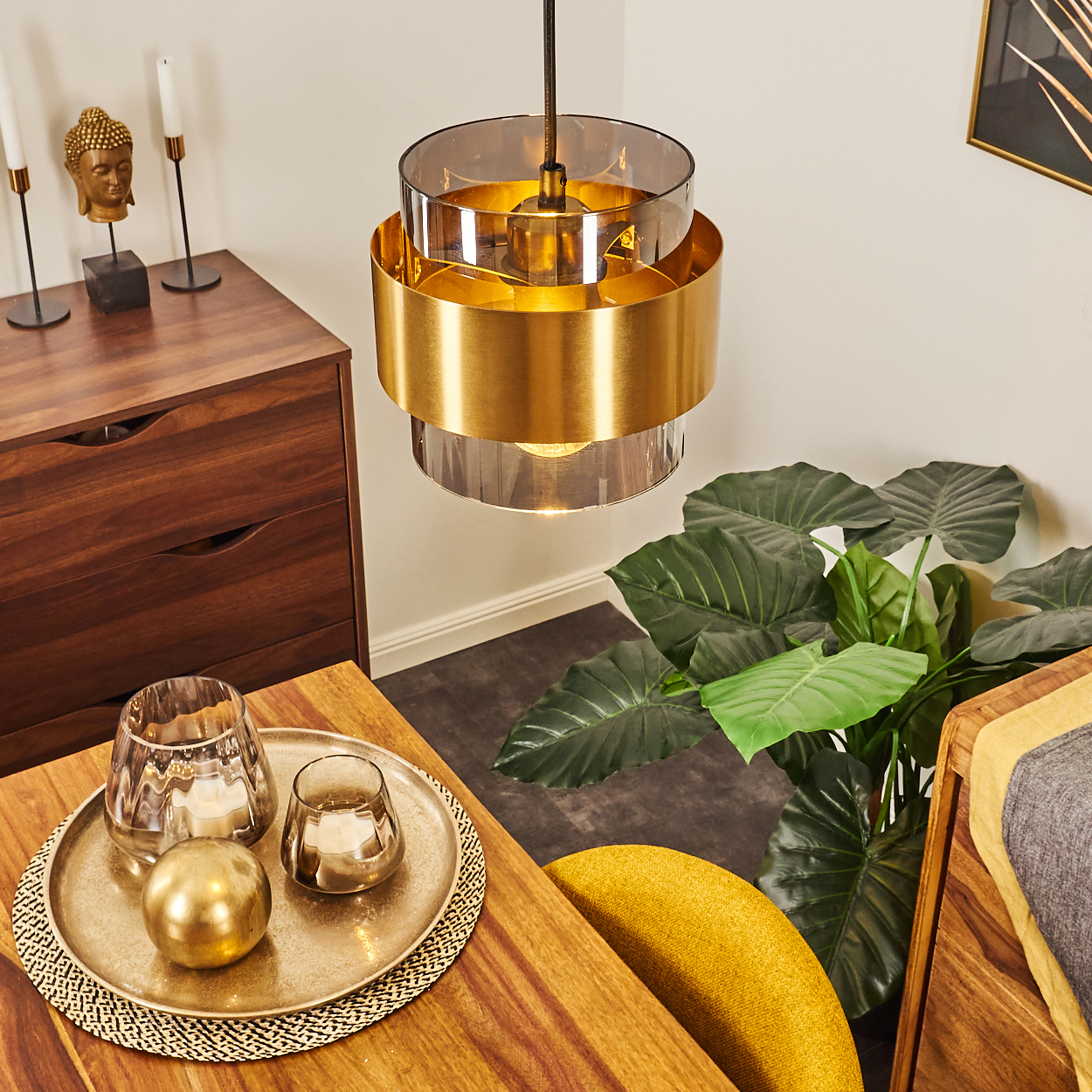 Eine moderne Innenszene. Über einem Holztisch mit Dekoration, einer Pflanze und einem gelben Stuhl hängt ein goldener Kronleuchter. Im Hintergrund ein Holzschrank.