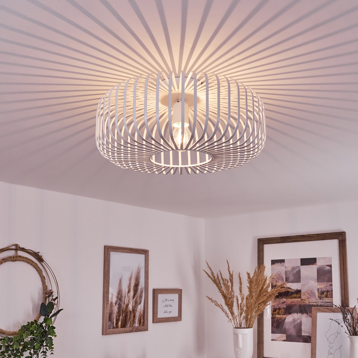 Eine weiße, kreisförmige Deckenlampe mit Lamellen wirft gestreifte Schatten. Darunter schmücken gerahmte Bilder und Vasen mit Pflanzen den Raum.
