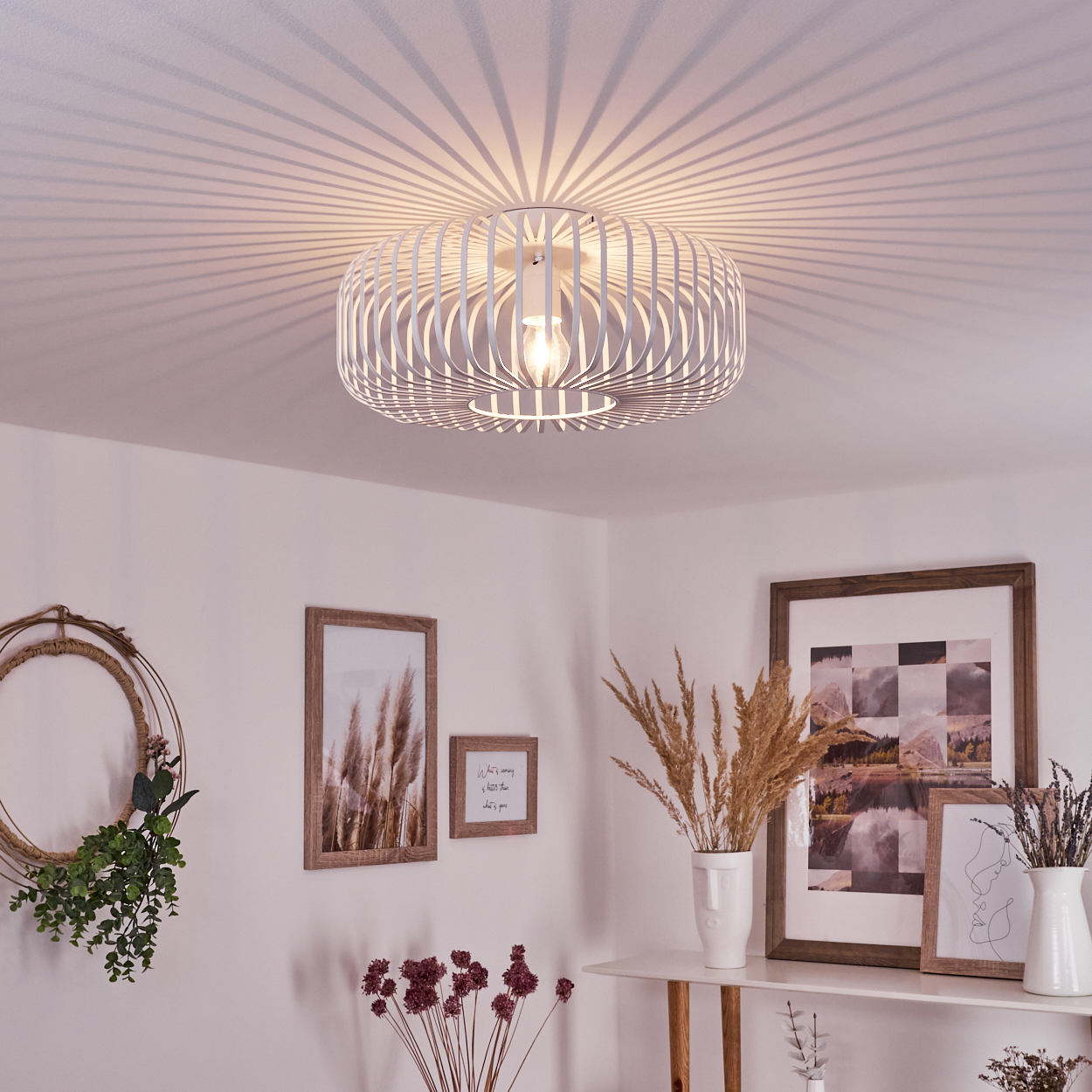 Eine weiße Deckenlampe wirft Schatten. Darunter befinden sich gerahmte Bilder, Vasen mit Pflanzen und ein dekorativer Kranz.