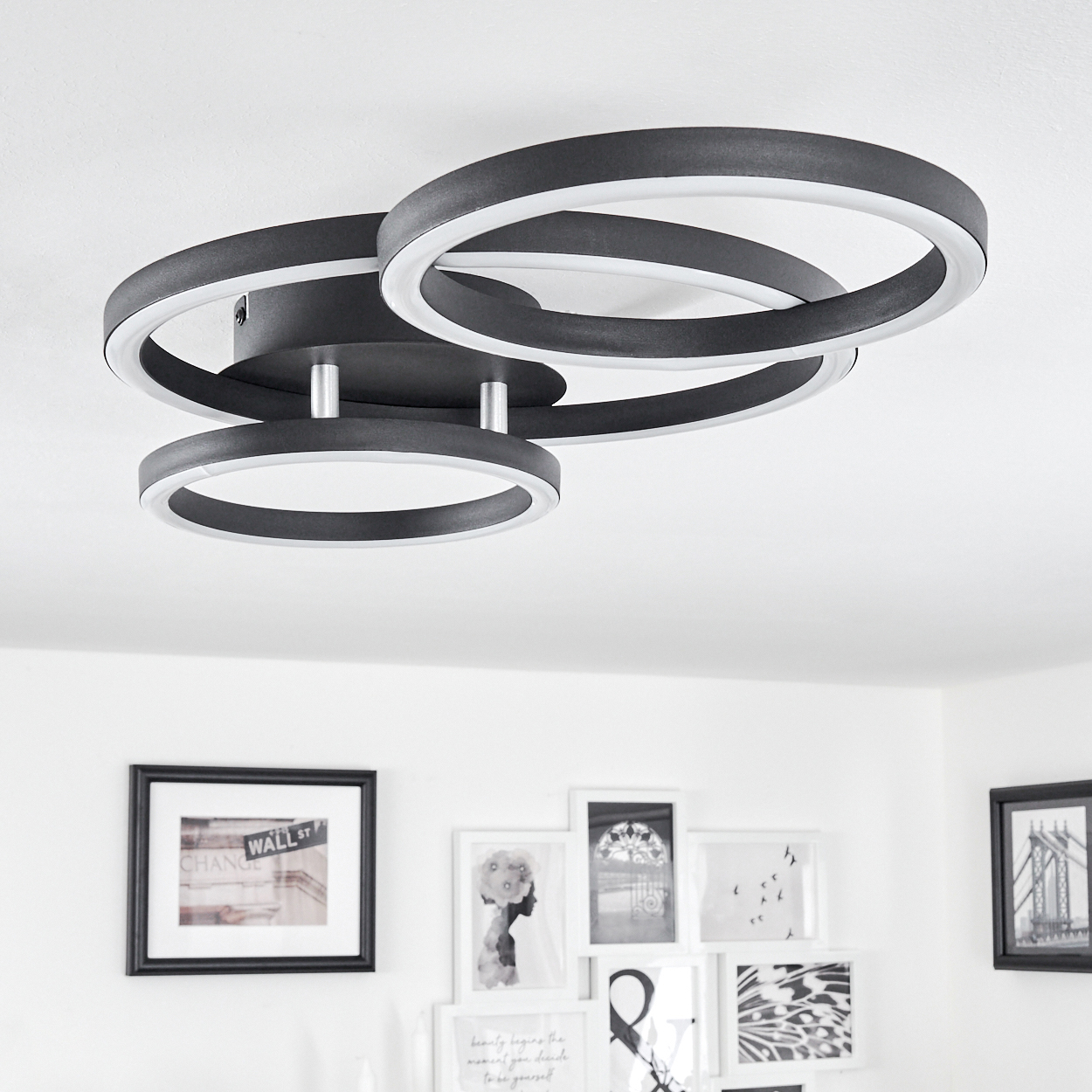 Eine moderne Deckenleuchte mit drei schwarz-weißen kreisförmigen Lichtern. Darunter befinden sich mehrere gerahmte Bilder an einer weißen Wand.