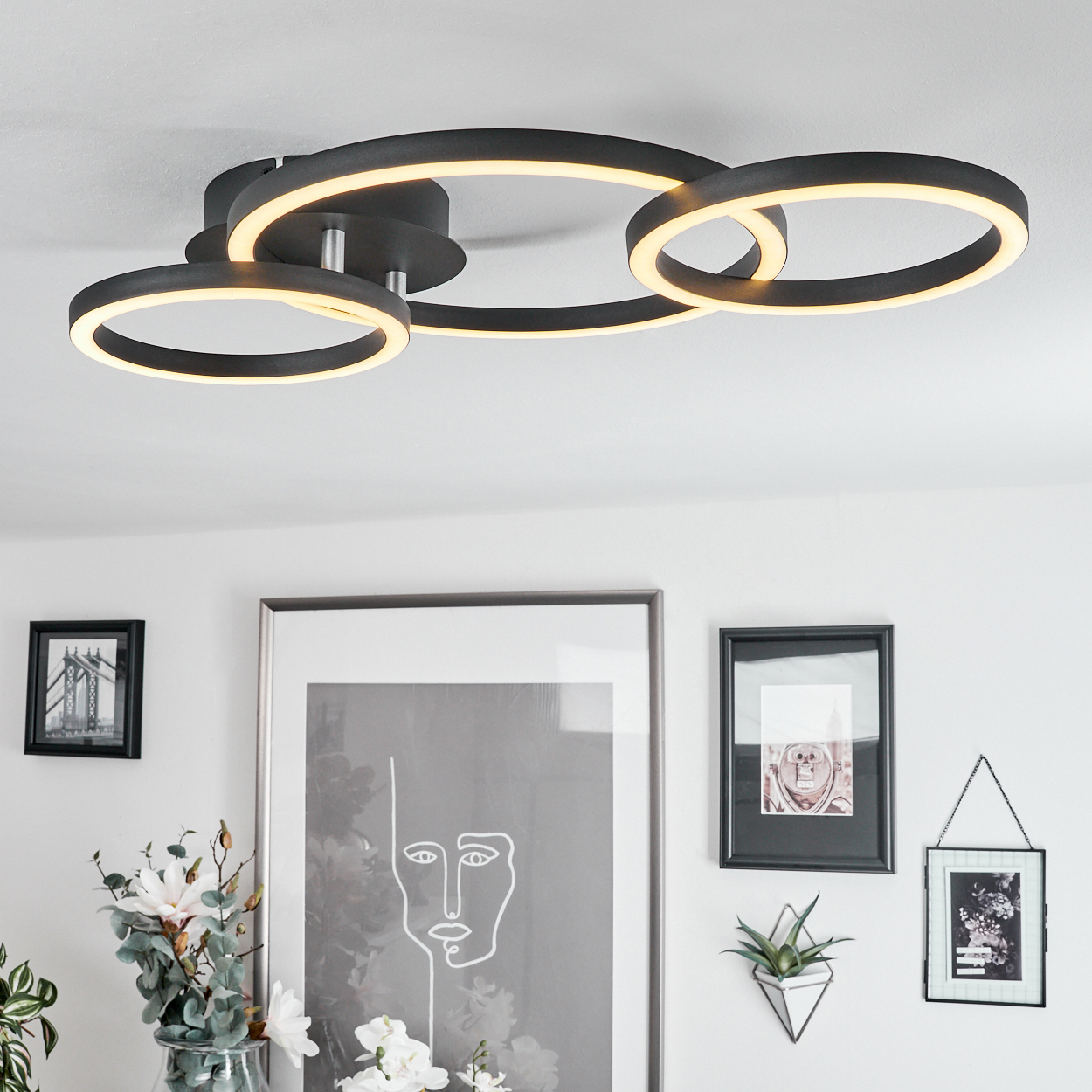 Moderne Deckenleuchte mit drei schwarzen Ringen, die warmes Licht aussenden. Darunter sind gerahmte Kunst und Dekoration an einer weißen Wand zu sehen.