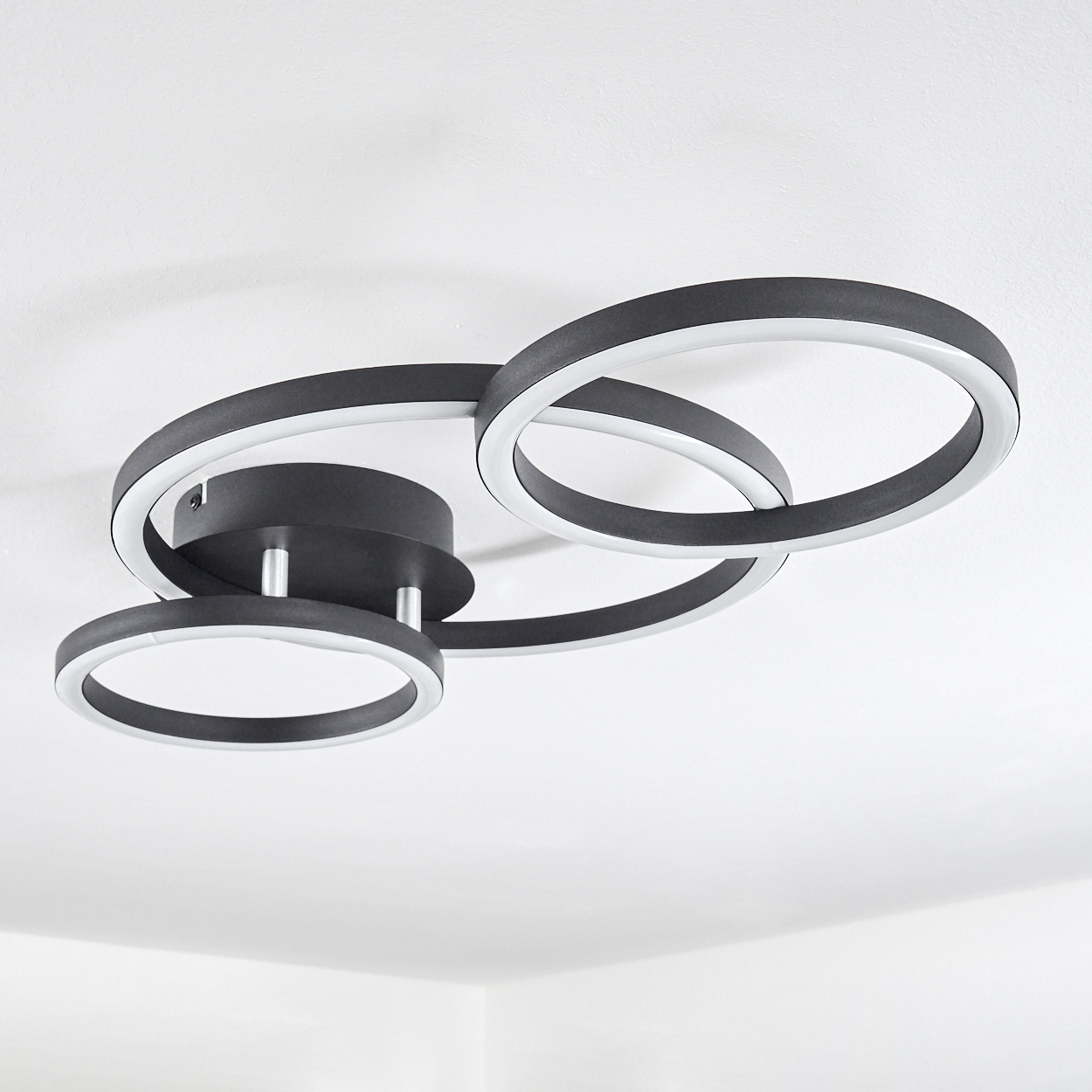Moderne Deckenleuchte. Sie hat drei miteinander verbundene, schwarze, ringförmige Lichter mit weißen Innenringen, vor einer weißen Decke.