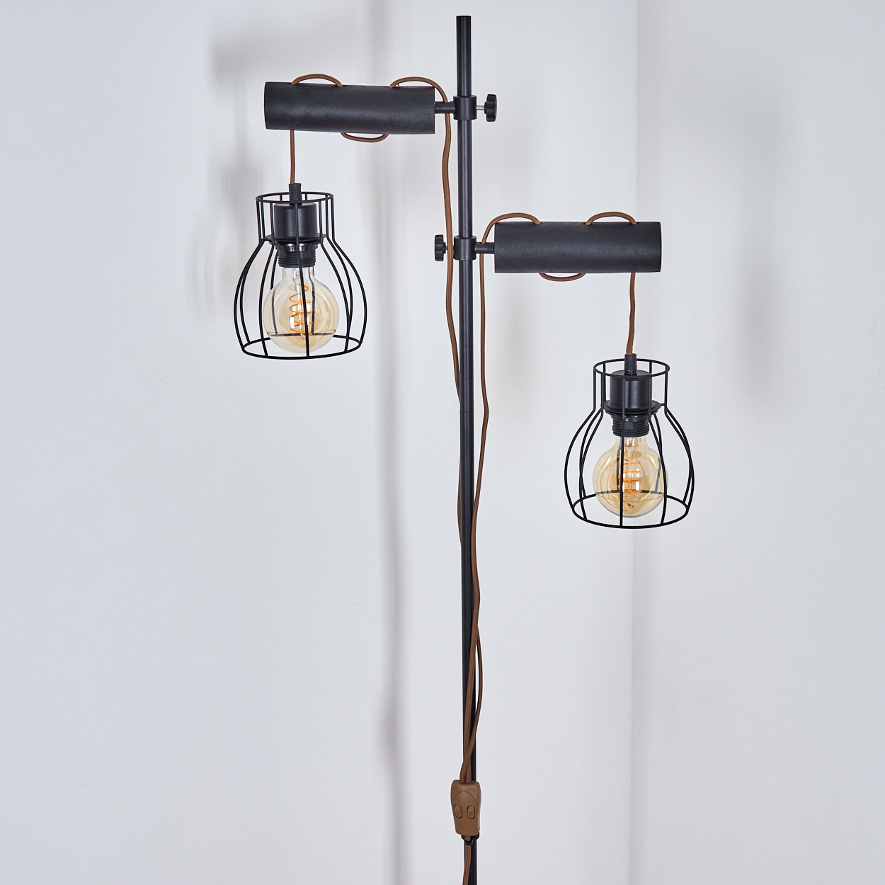 Eine schwarze Metall-Stehlampe mit zwei käfigartigen Glühbirnen. Braune Drähte und ein Schalter sind sichtbar. Der Hintergrund ist eine weiße Eckwand.