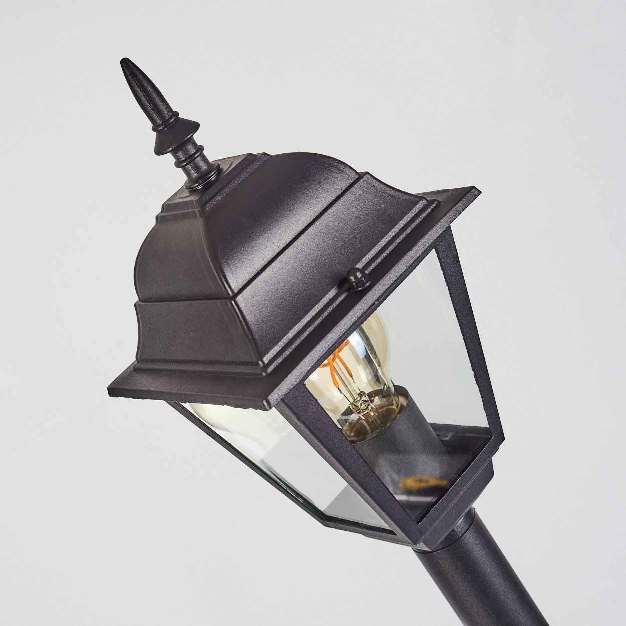 Eine schwarze Außenlampe mit spitzem Aufsatz und Glasscheiben. Eine Edison-Glühbirne ist sichtbar. Der Hintergrund ist weiß.