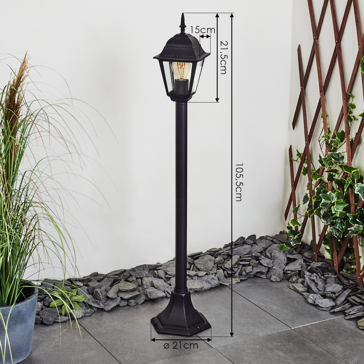 Schwarze Außenlampe mit Glühbirne, die auf einer gefliesten Oberfläche steht. Sie befindet sich neben einer Pflanze, einem Holzgitter und Steinen.