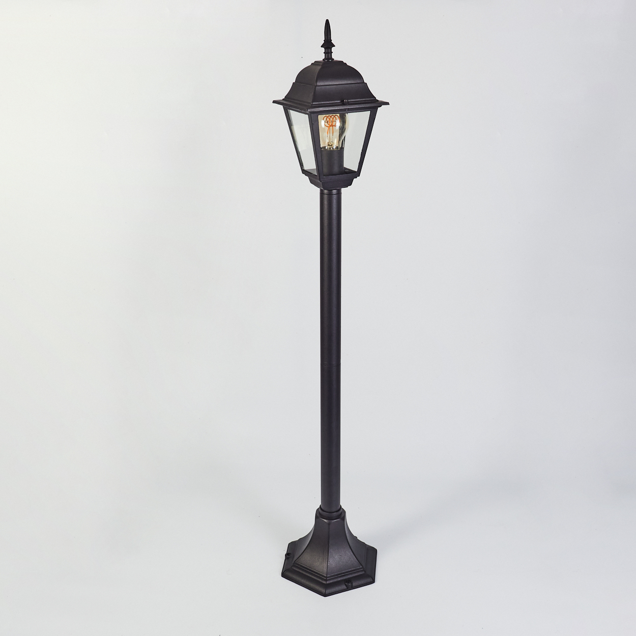 Eine schwarze Außenlaterne steht auf weißem Hintergrund. Die Lampe hat ein Glasgehäuse mit einer leuchtenden Glühbirne.