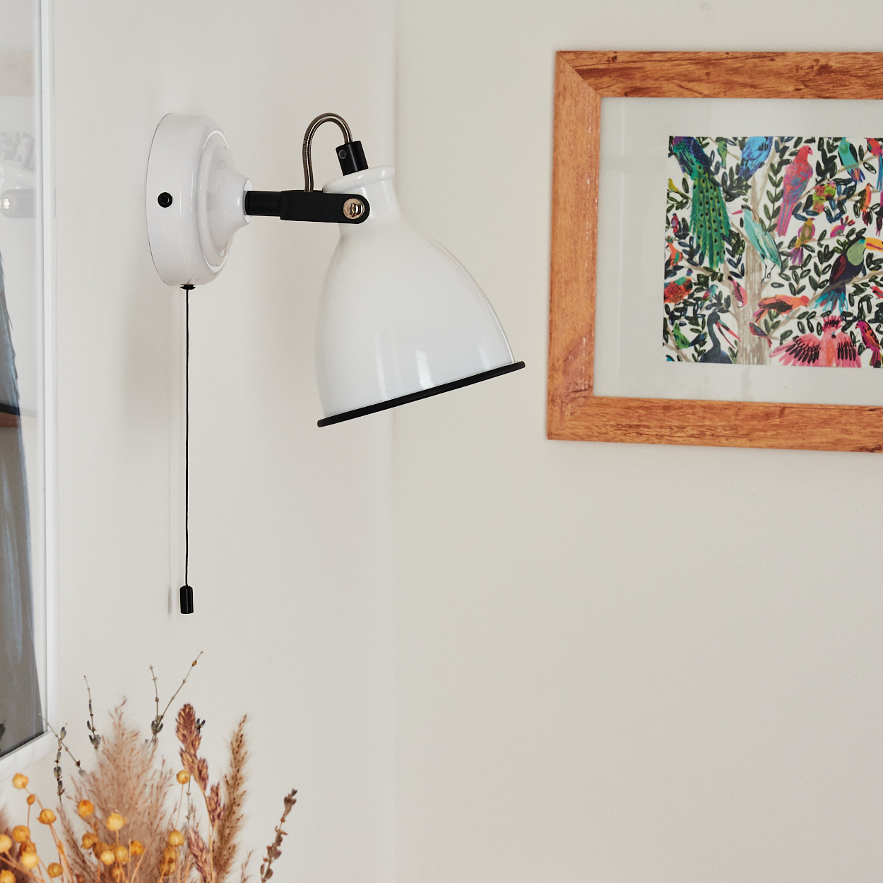 Weiße Wandlampe mit schwarzen Details, ein gerahmtes Vogelbild und Trockenblumen in einer Ecke an einer weißen Wand.