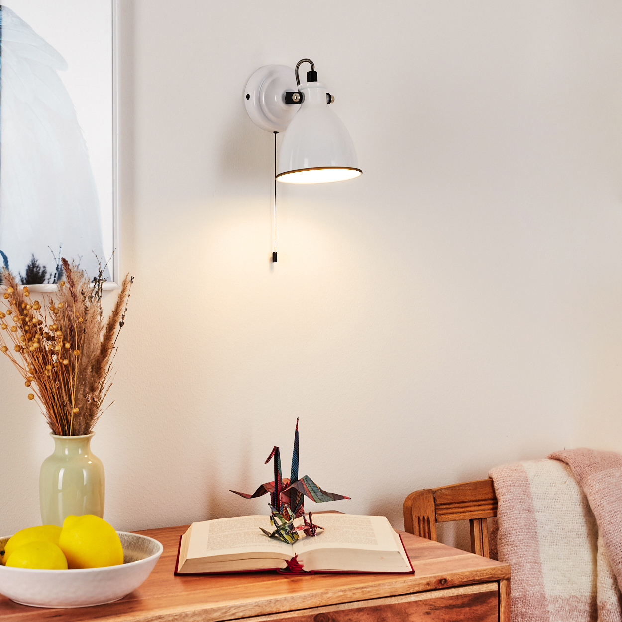 Eine weiße Wandlampe beleuchtet einen Holztisch mit Buch, Origami-Kranich, Vase mit Blumen, Zitronen und Decke.