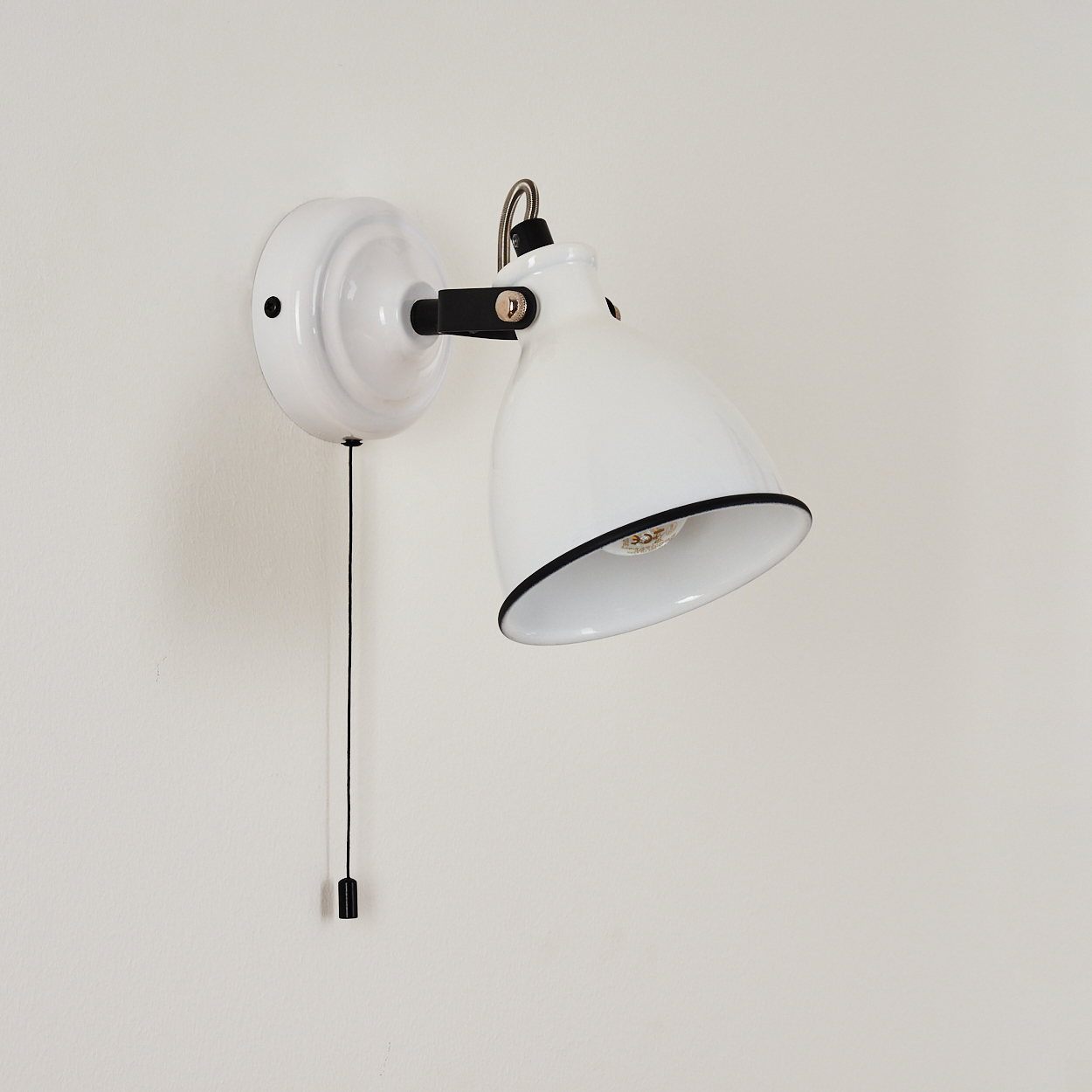 Eine weiße Wandleuchte mit Zugkordel. Die Lampe hat einen schwarzen Rand und eine schwarze Halterung. Sie befindet sich an einer weißen Wand.
