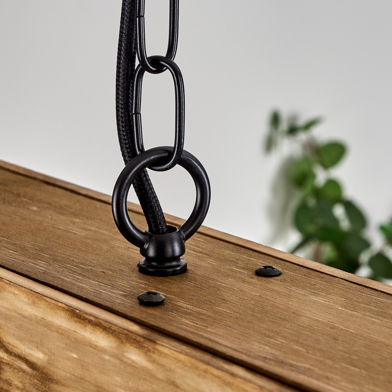 Nahaufnahme eines Holzbretts mit einem schwarzen Metallring und einer Kette. Ein schwarzes Kabel führt durch den Ring. Zwei Schrauben sind sichtbar.
