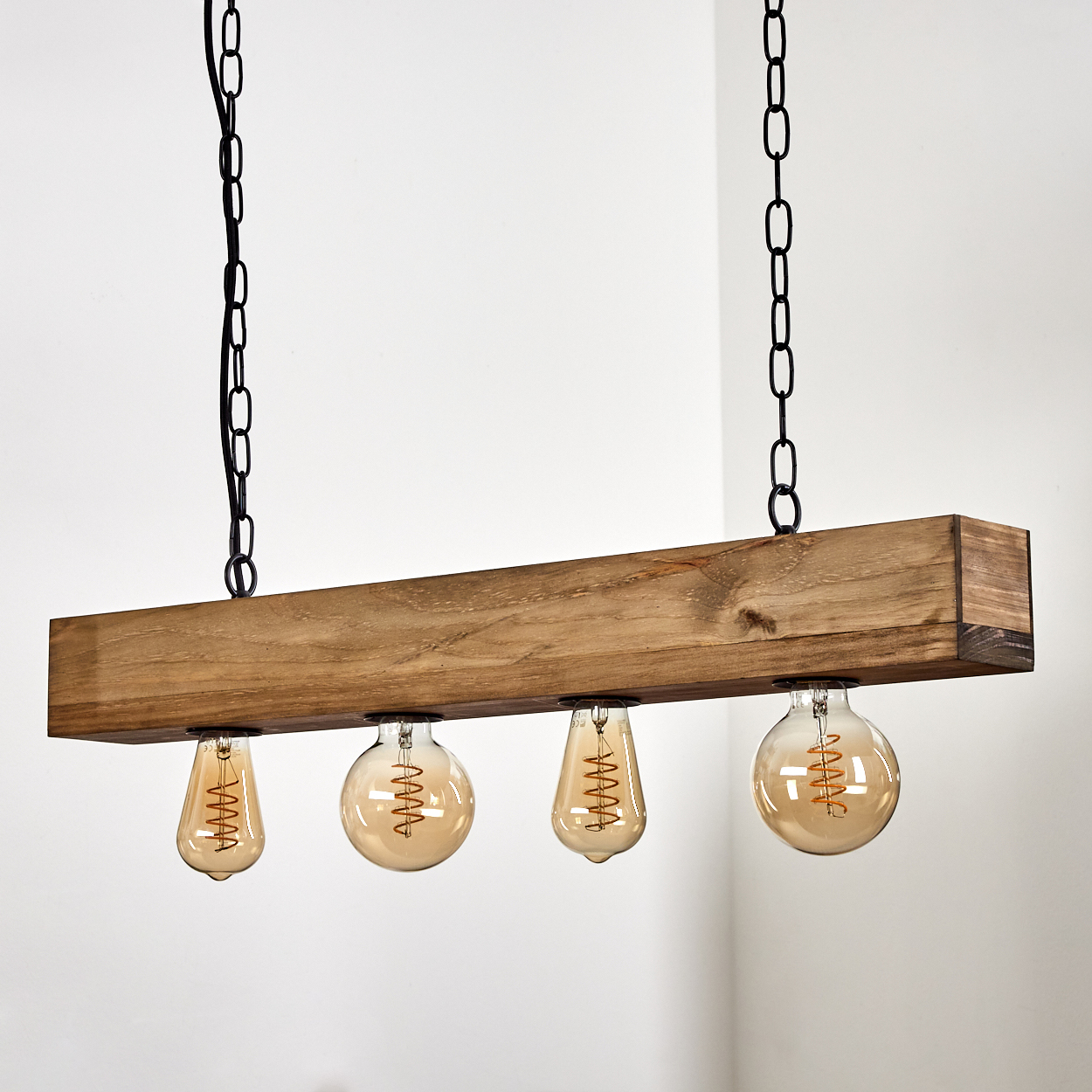 Ein Holzbalken mit vier Glühbirnen hängt an schwarzen Ketten. Die Glühbirnen sind warmorangefarben und der Hintergrund ist eine weiße Wand.