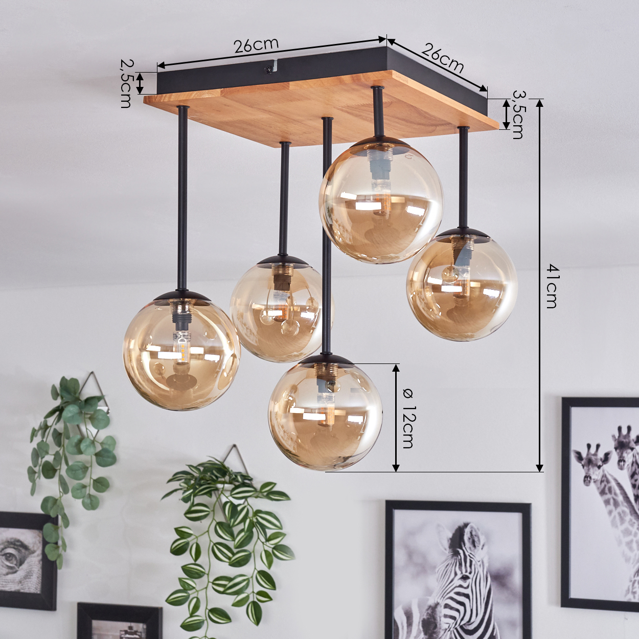 Eine Deckenlampe mit fünf bernsteinfarbenen Glaskugeln. Holzsockel mit schwarzen Metallstützen. Pflanzen und gerahmte Bilder im Hintergrund.