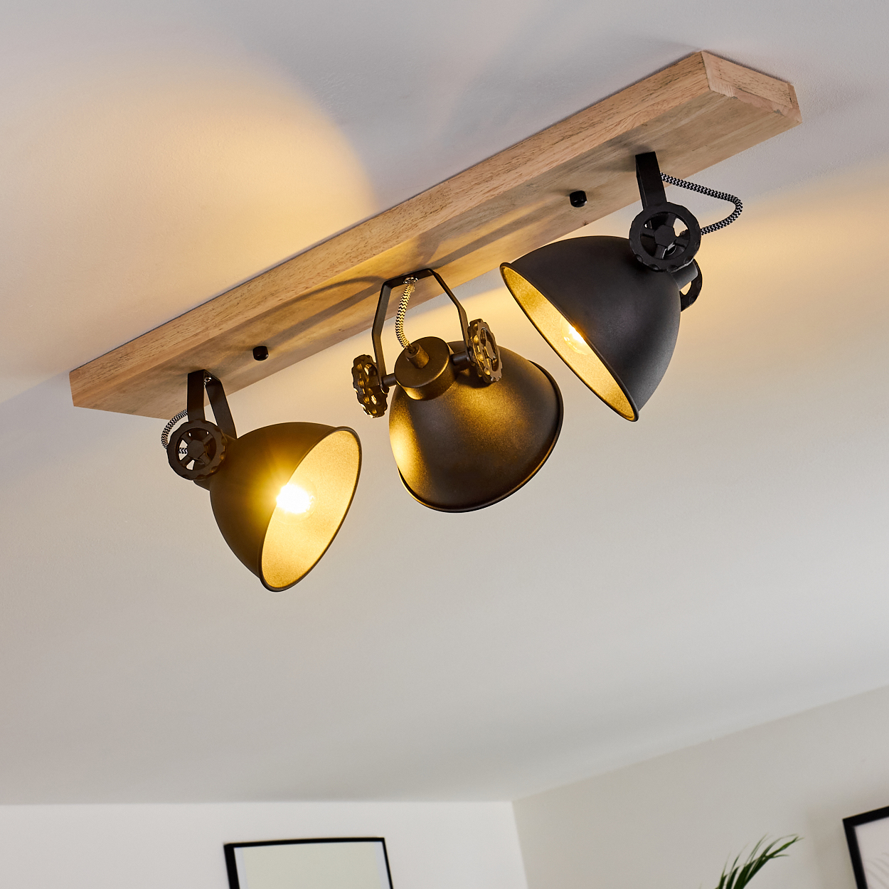 Eine Deckenstrahler-Leuchte mit drei schwarzen Lampen auf einem Holzbalken, die nach unten leuchten. Der Hintergrund ist eine weiße Decke.