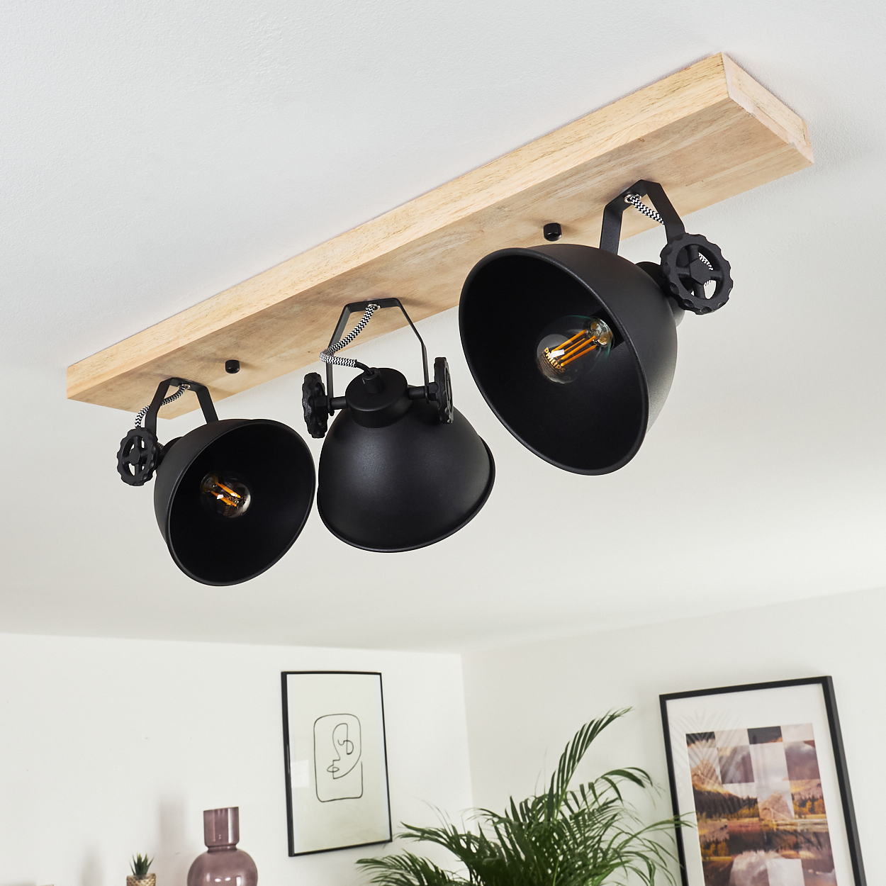 Eine schwarze Strahler-Leuchte mit drei Lichtern hängt an einem Holzbalken. Der Hintergrund zeigt eine weiße Decke und Wand mit Dekor.