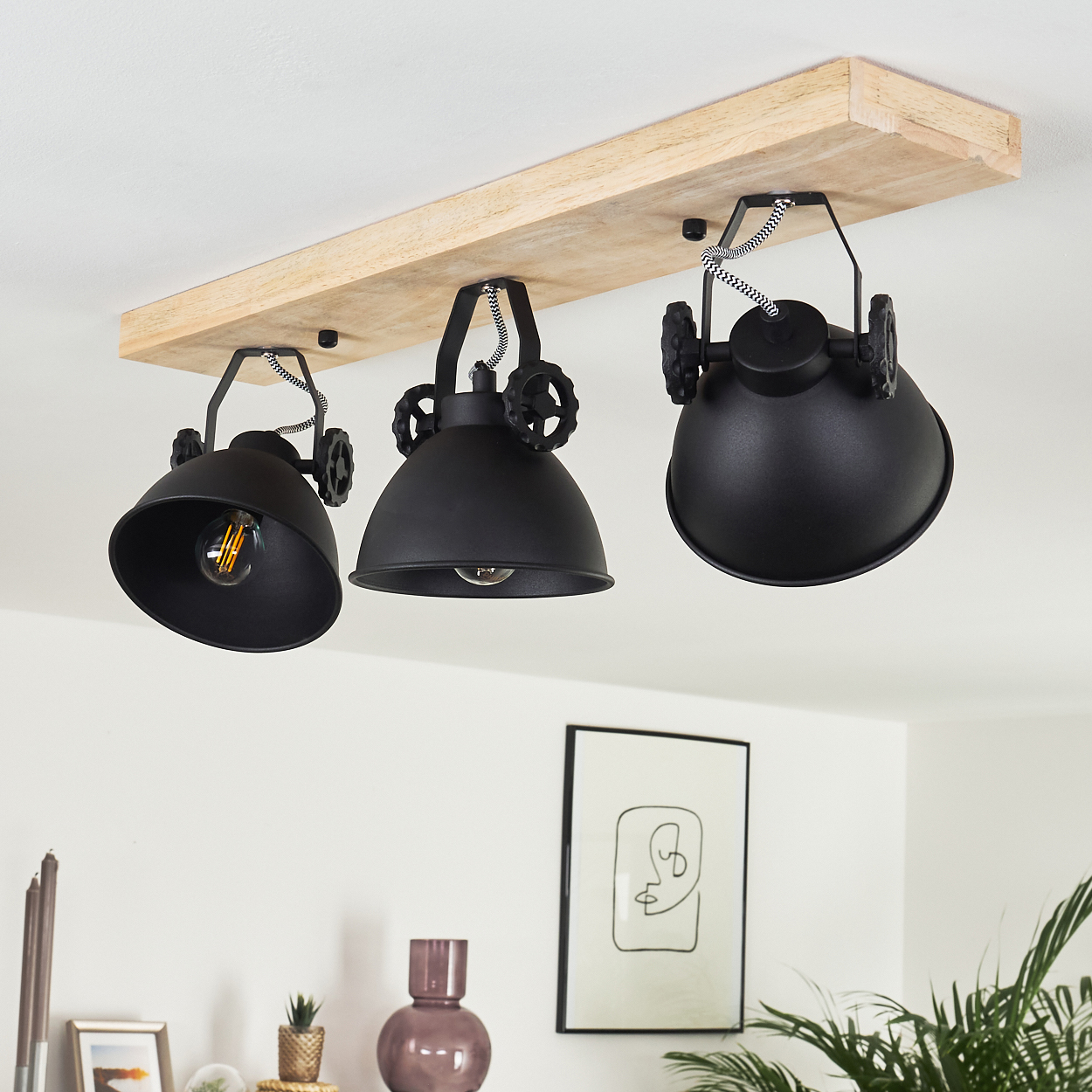 Drei schwarze Hängelampen mit sichtbaren Glühbirnen hängen an einem Holzbalken. Ein Bild und Pflanzen sind unten sichtbar.
