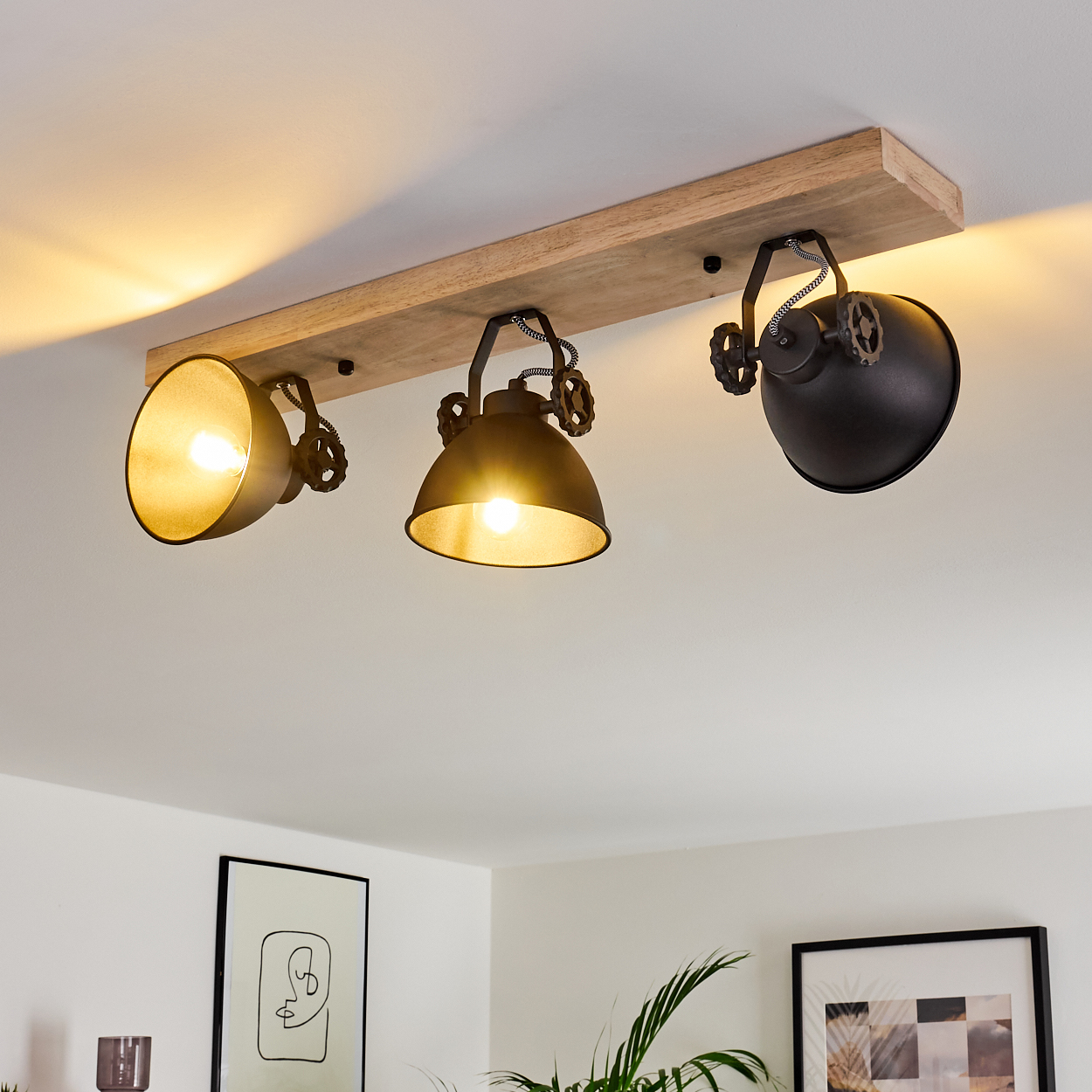 Deckenleuchte mit drei schwarzen Lampen an einem Holzbalken. Die Lichter sind an. Im Hintergrund ist ein Raum mit Kunst und Pflanzen zu sehen.