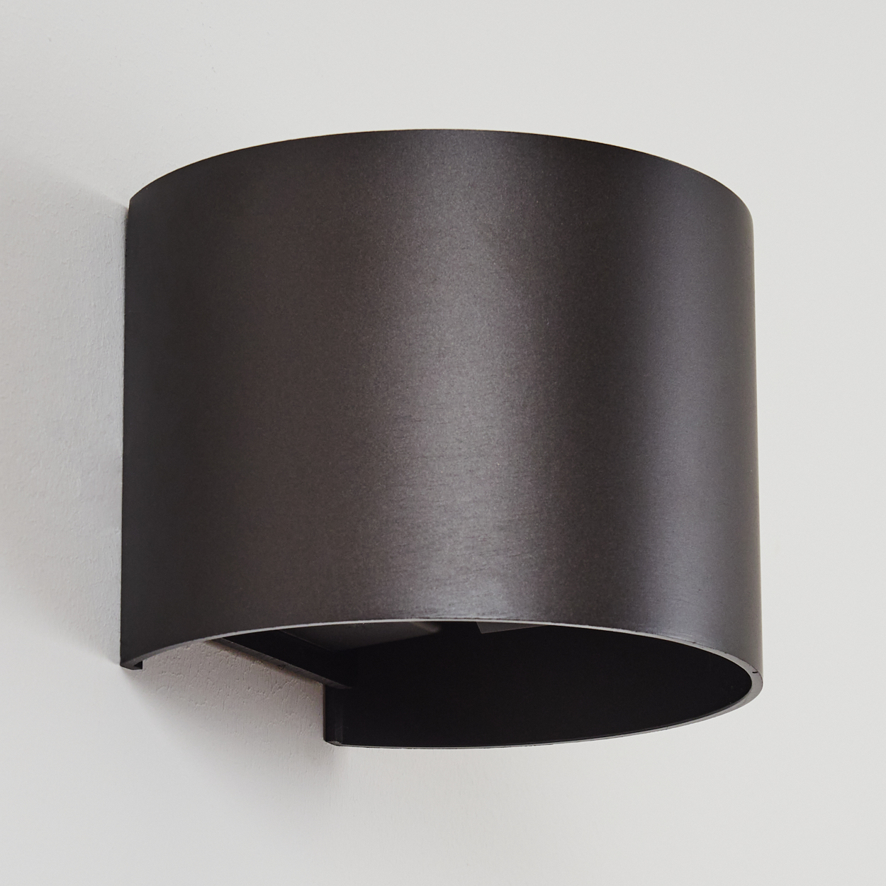 Eine dunkelgraue, halbkreisförmige Wandleuchte ist an einer weißen Wand befestigt. Die Lampe hat ein gebogenes, zylindrisches Design mit offenem Boden.