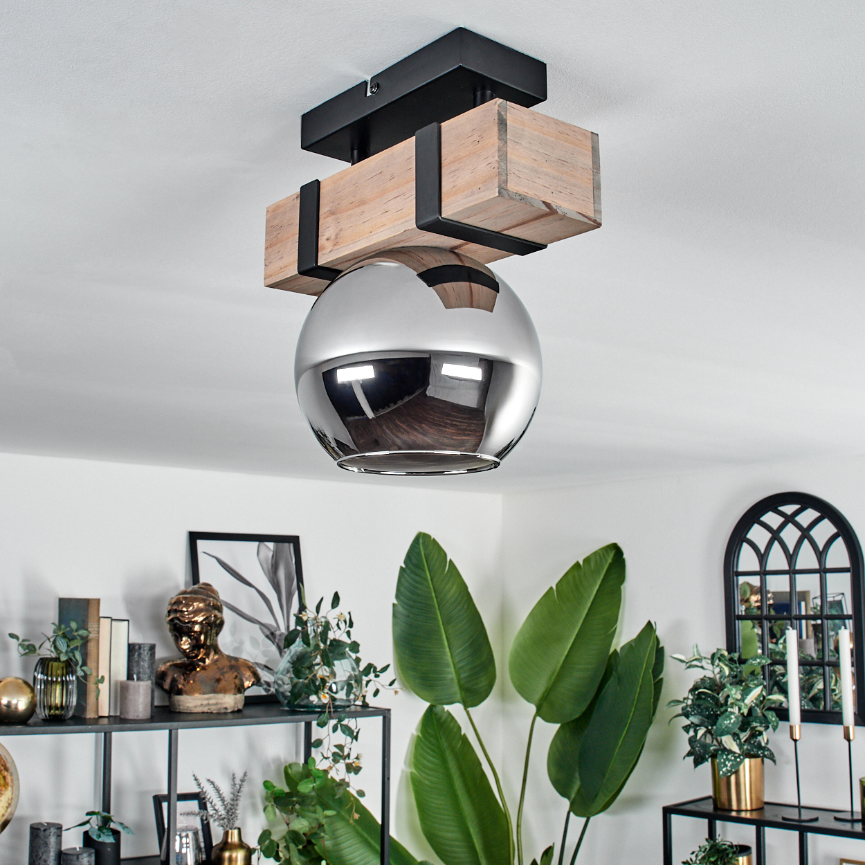 Eine moderne Deckenlampe mit einer Chromkugel. Holzbalken und schwarze Metallakzente. Grüne Pflanzen und Dekorationsartikel im Hintergrund.
