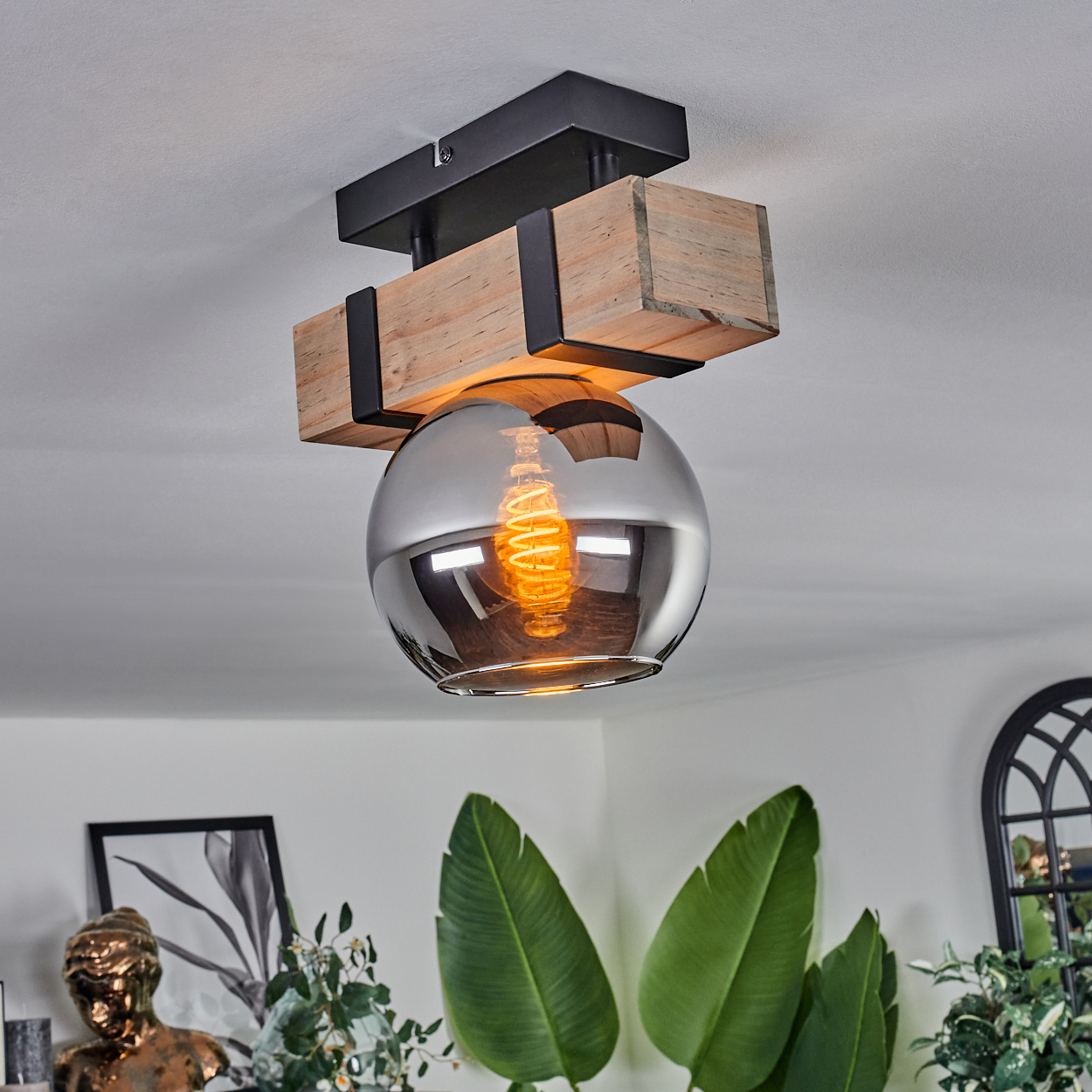 Eine Deckenleuchte mit Holzbalken, Glaskugel und Edison-Glühbirne. Grüne Pflanzen und Dekoration im Hintergrund.
