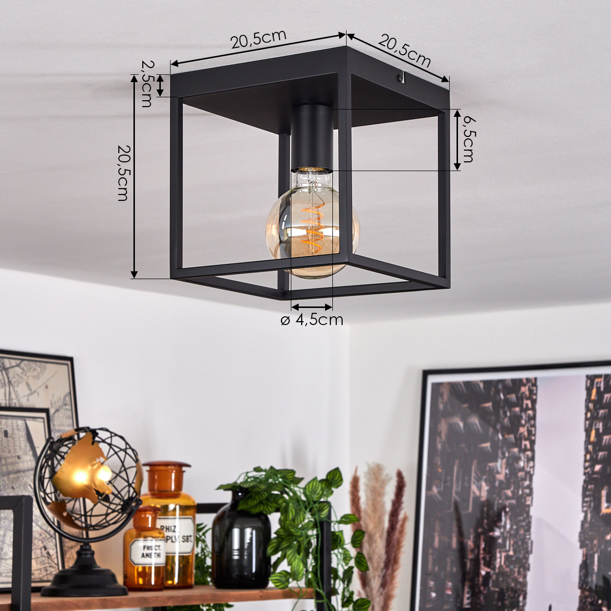 Schwarze quadratische Deckenlampe mit runder Glühbirne. Abmessungen sind beschriftet. Darunter ein Regal mit Dekorationen und Pflanzen.