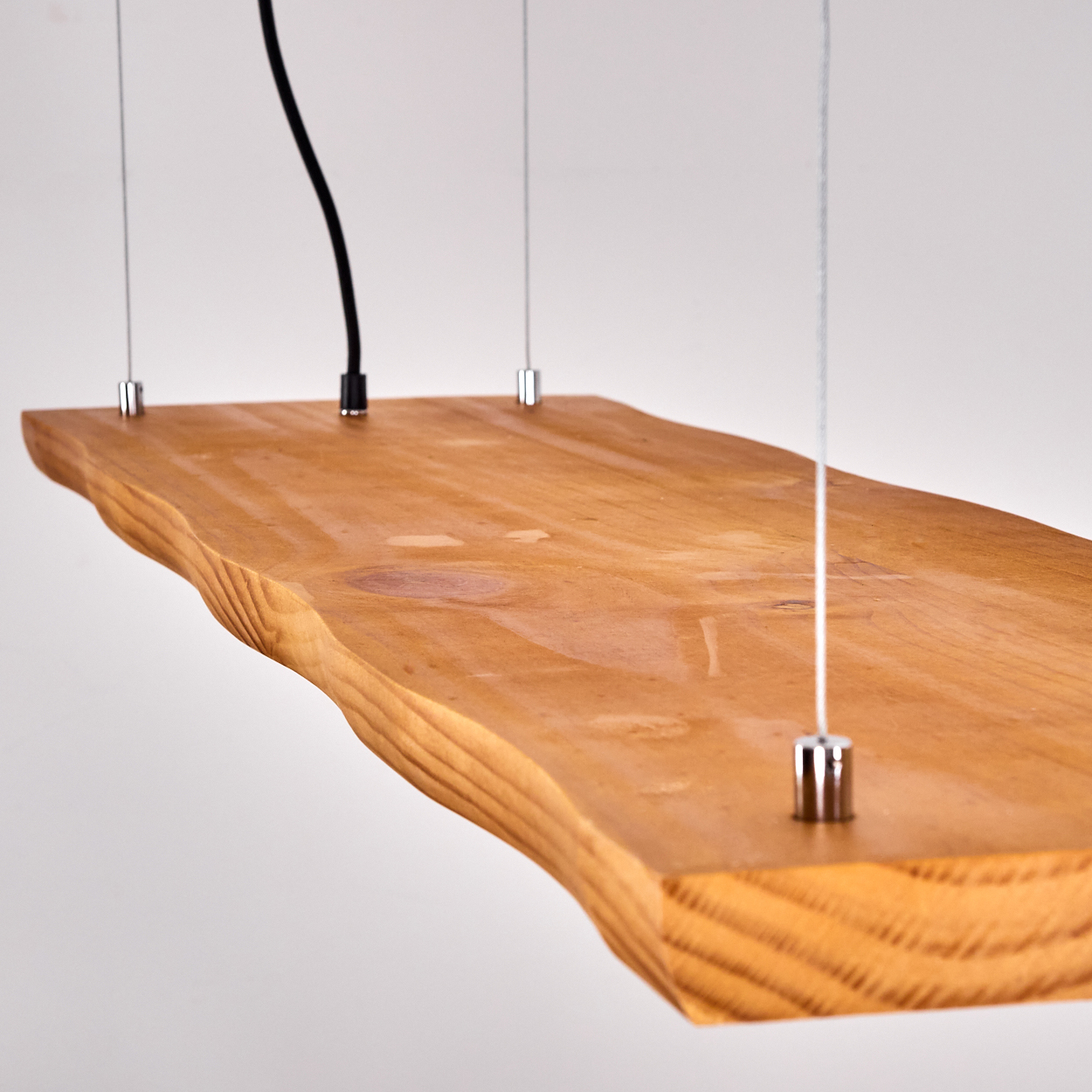 Ein Holzregal, das an Drähten aufgehängt ist. Es hat eine natürliche Kante und wird von oben beleuchtet. Ein schwarzes Kabel hängt vom Regal.