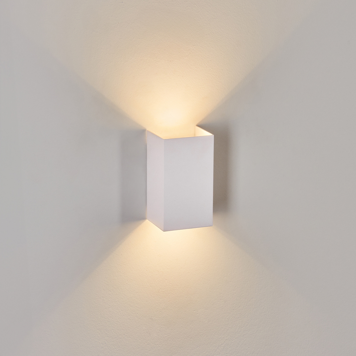 Eine weiße, eckige Wandleuchte ist an einer hellgrauen Wand montiert. Die Lampe strahlt warmes Licht aus und wirft Schatten nach oben und unten.