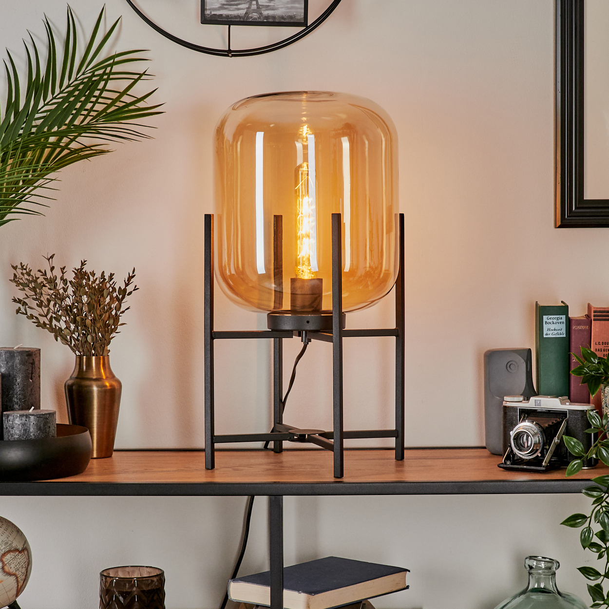 Eine beleuchtete orangefarbene Glaslampe auf einem schwarzen Metallständer steht auf einem Holzregal. Weitere Gegenstände sind Kerzen, Bücher und eine Kamera.