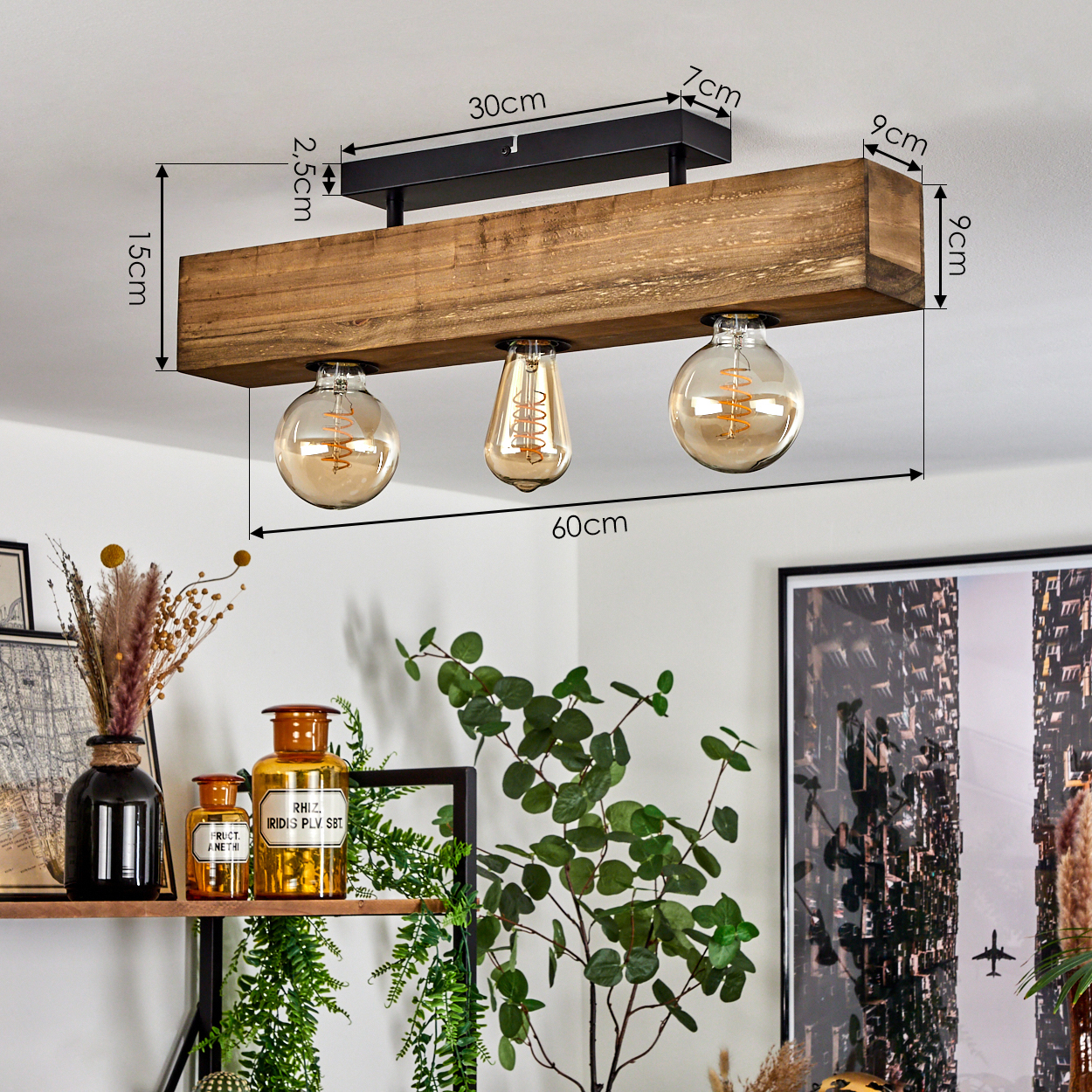 Holzdeckenlampe mit drei Glühbirnen. Darunter Pflanzen und dekorative Flaschen. Im Hintergrund ein gerahmtes Bild.