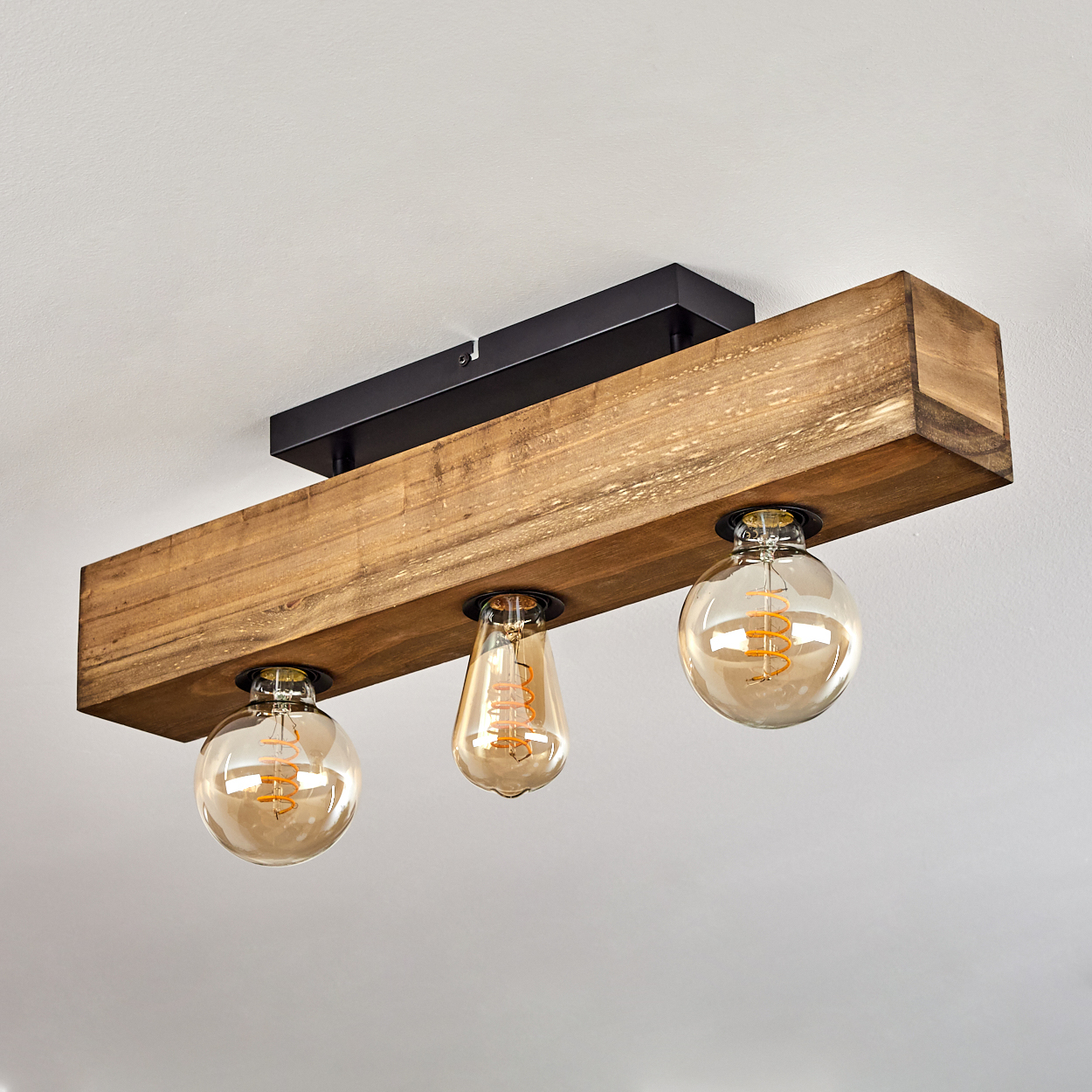 Eine Deckenleuchte aus Holzbalken mit drei sichtbaren Glühbirnen. Zwei sind rund, eine tropfenförmig. Die Decke ist weiß.